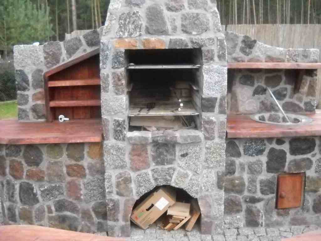 Kamienny grill ogrodowy z paleniskiem, rusztem, drewnianymi półkami i schowkiem na akcesoria, otoczony kostką brukową i zielenią.