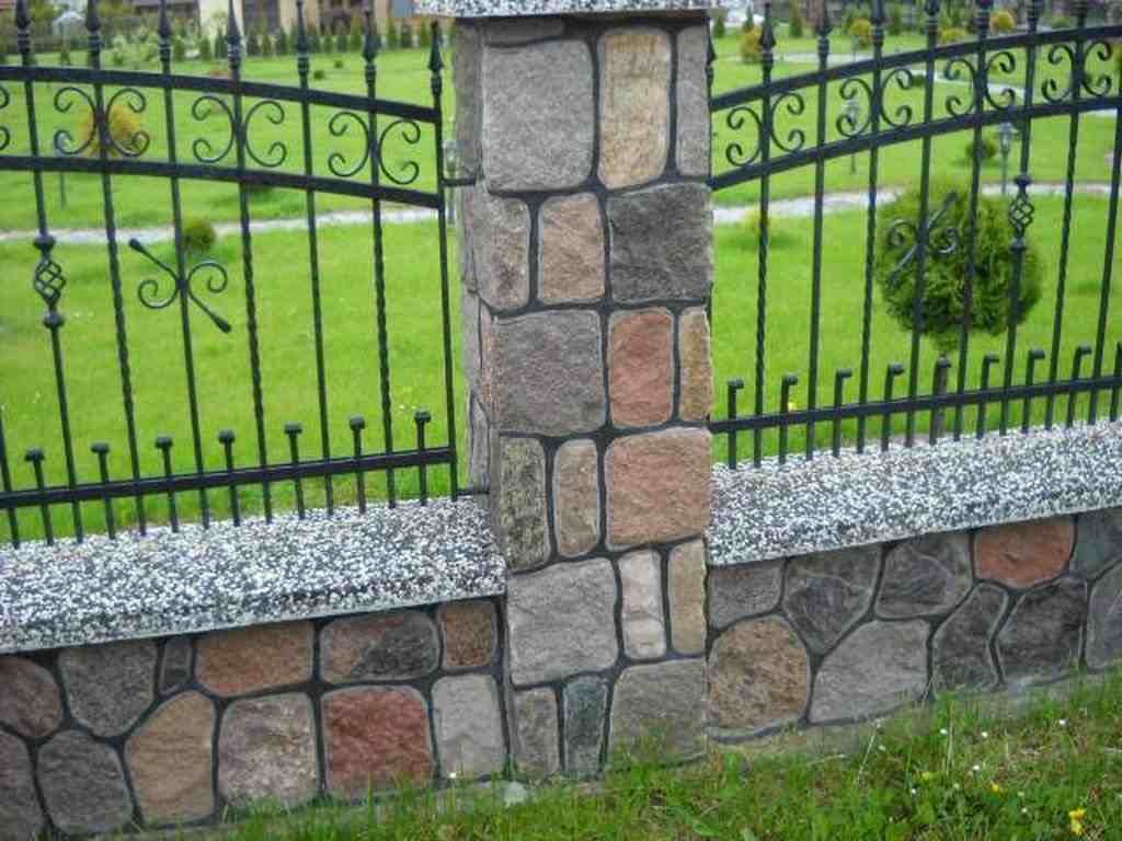 Żelazny płot z kamienną podmurówką i słupem z nieregularnych kamieni polnych w odcieniach szarości i brązu, na tle zielonego trawnika.