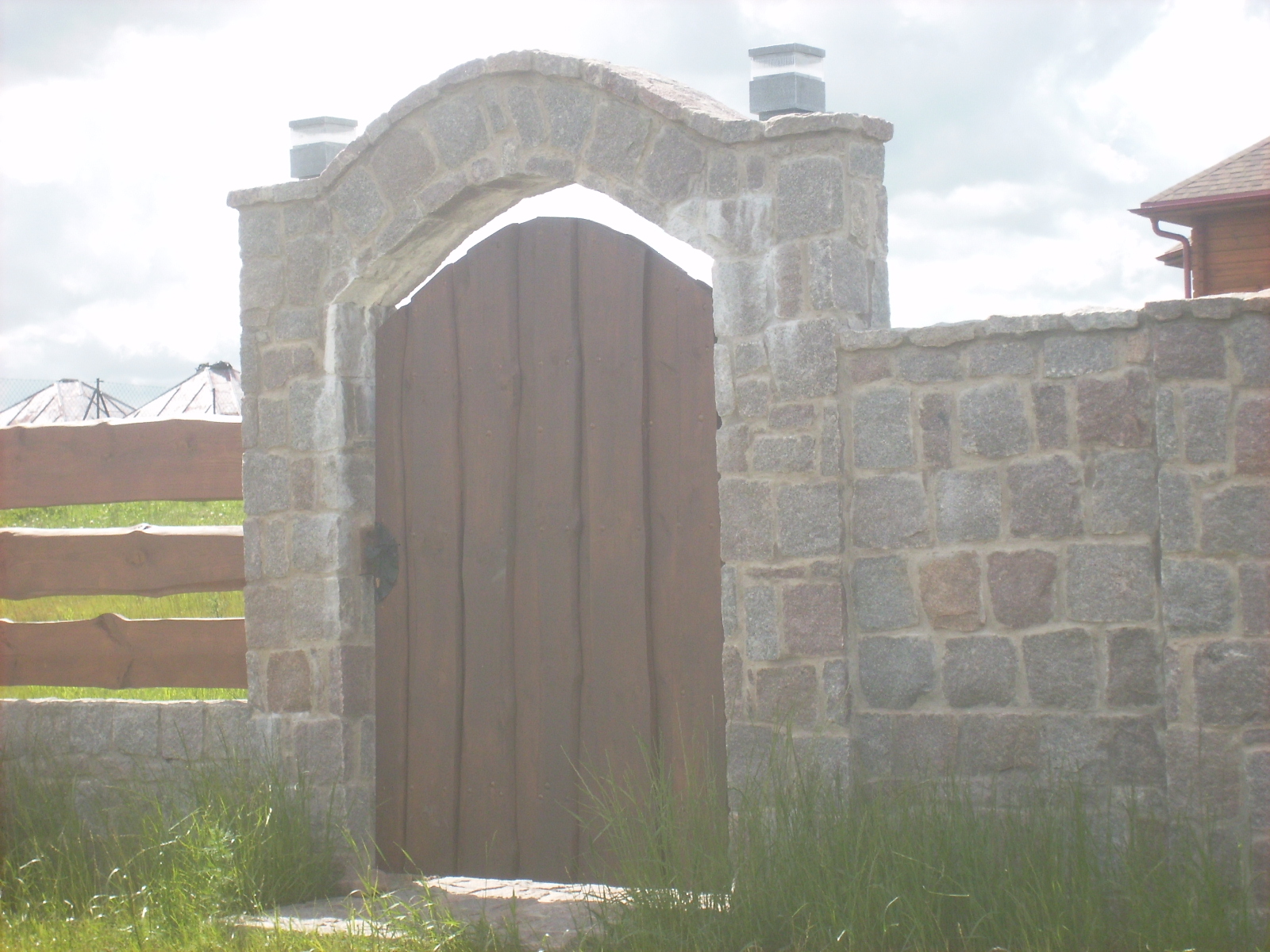 Wejście na posesję z drewnianą bramą w kamiennej obudowie z łukiem i lampami na szczycie, otoczone niskim murem z kamienia polnego.