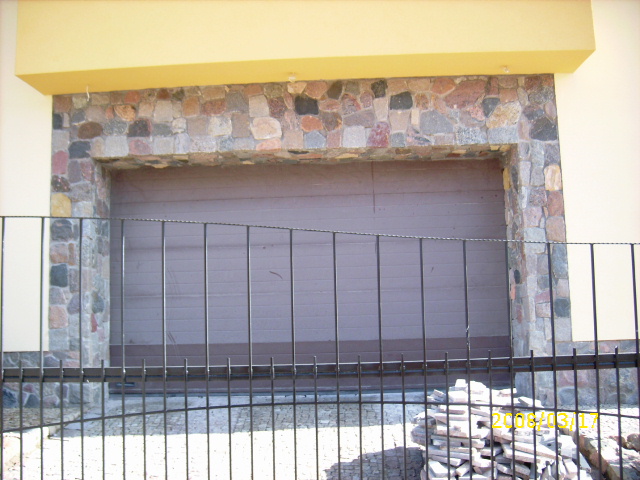 Brama garażowa obłożona nieregularnym kamieniem elewacyjnym w odcieniach szarości, brązu i różu, widoczna zza metalowego ogrodzenia.