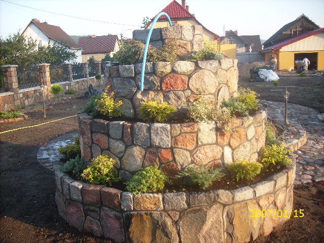 Trzykondygnacyjna fontanna ogrodowa z nieregularnych kamieni polnych, obsadzona zielenią, z niebieską rurką doprowadzającą wodę, na tle ogrodu w trakcie prac wykończeniowych.