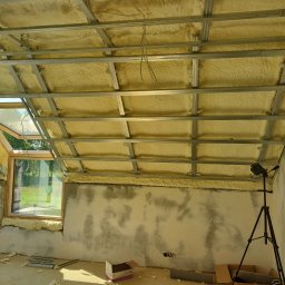 Serhii Ivanchuk - Poddasze w trakcie ocieplania pianą poliuretanową, widoczna konstrukcja metalowa sufitu, okno dachowe, ślady po pracach na ścianach, laser krzyżowy na statywie, resztki piany na podłodze.