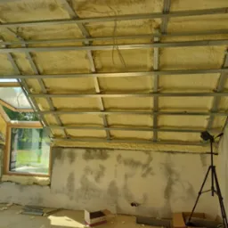 Poddasze w trakcie ocieplania pianą poliuretanową, widoczna konstrukcja metalowa sufitu, okno dachowe, ślady po pracach na ścianach, laser krzyżowy na statywie, resztki piany na podłodze.