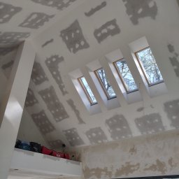 Serhii Ivanchuk - Poddasze w trakcie przygotowania ścian i sufitu do dalszych prac wykończeniowych, widoczne łaty szpachli na płytach gipsowo-kartonowych, cztery okna dachowe w rzędzie oraz narzędzia na antresoli.
