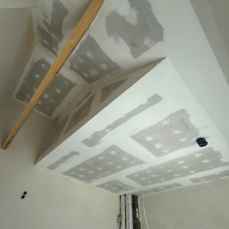 Serhii Ivanchuk - Sufit podwieszany z płyt karton-gips na etapie szpachlowania, widoczne łączenia płyt. Fragment drewnianej listwy na suficie. Instalacja elektryczna i rury w tle.