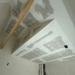 Sufit podwieszany z płyt karton-gips na etapie szpachlowania, widoczne łączenia płyt. Fragment drewnianej listwy na suficie. Instalacja elektryczna i rury w tle.