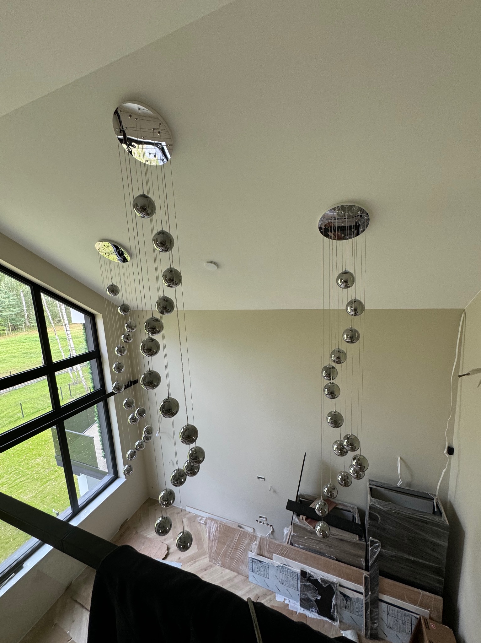 Dwa nowoczesne żyrandole kaskadowe z chromowanymi kulami zawieszonymi na cienkich linkach, widoczne na tle białych ścian i dużego okna z widokiem na zieleń, podłoga zabezpieczona folią, widoczne...