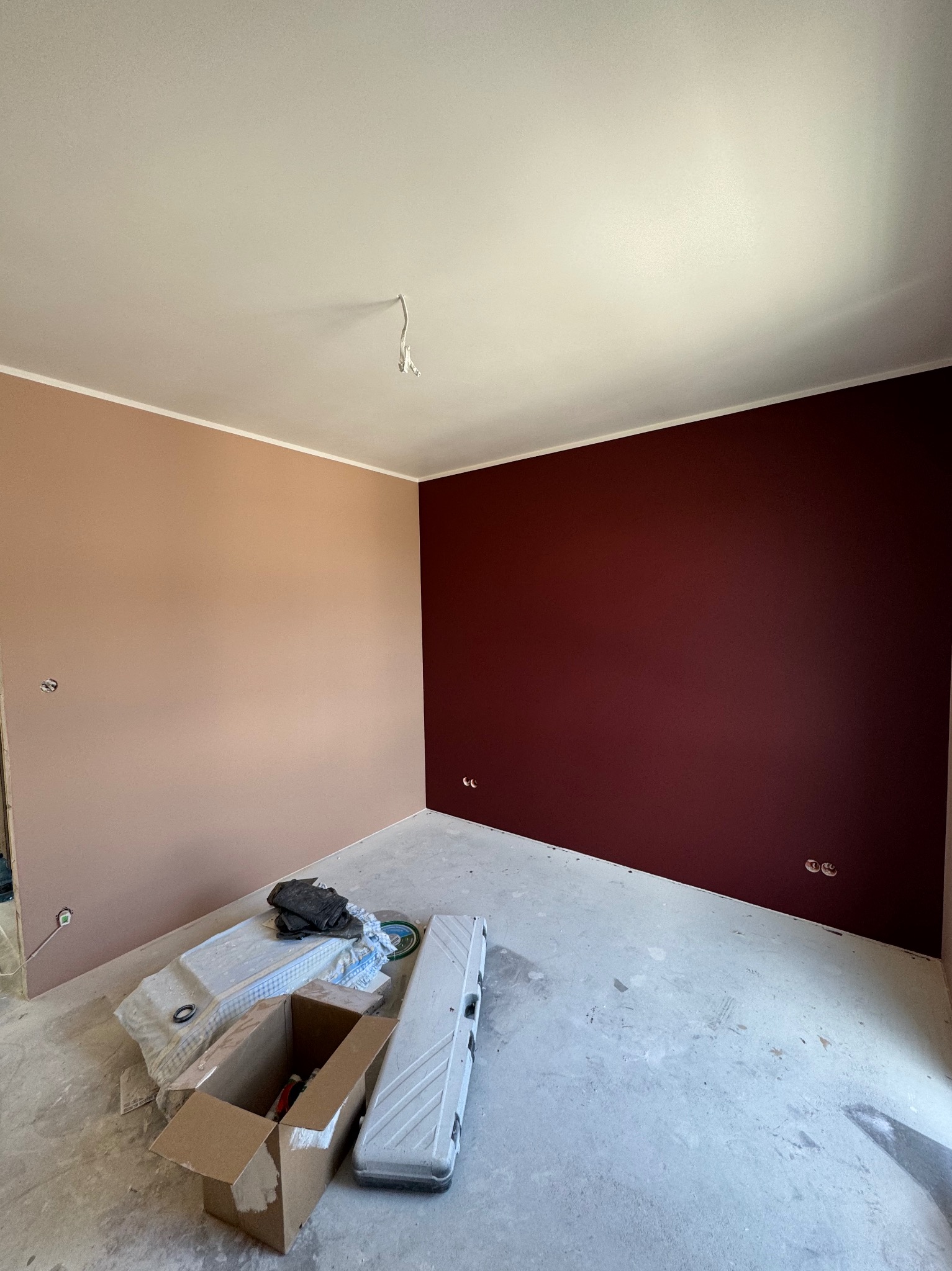 Pokój po malowaniu ścian na dwa kolory: beżowy i bordowy. Widoczne gniazdka elektryczne, narzędzia malarskie w kartonie i biała listwa przy suficie.
