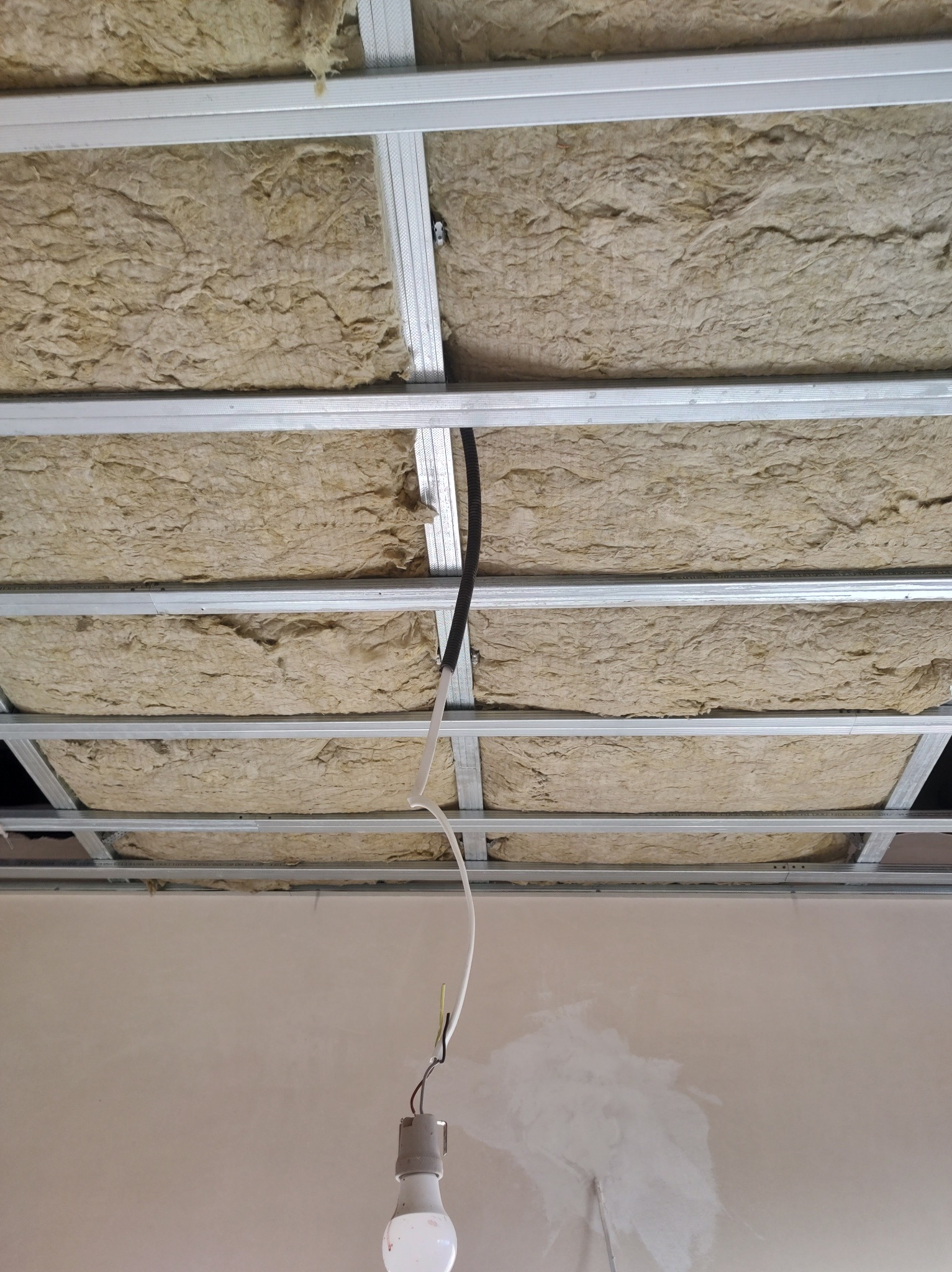 Poddasze z widoczną izolacją z wełny mineralnej pomiędzy metalowymi profilami sufitu podwieszanego, wystające przewody elektryczne z żarówką, ślady po szpachlowaniu na ścianie.