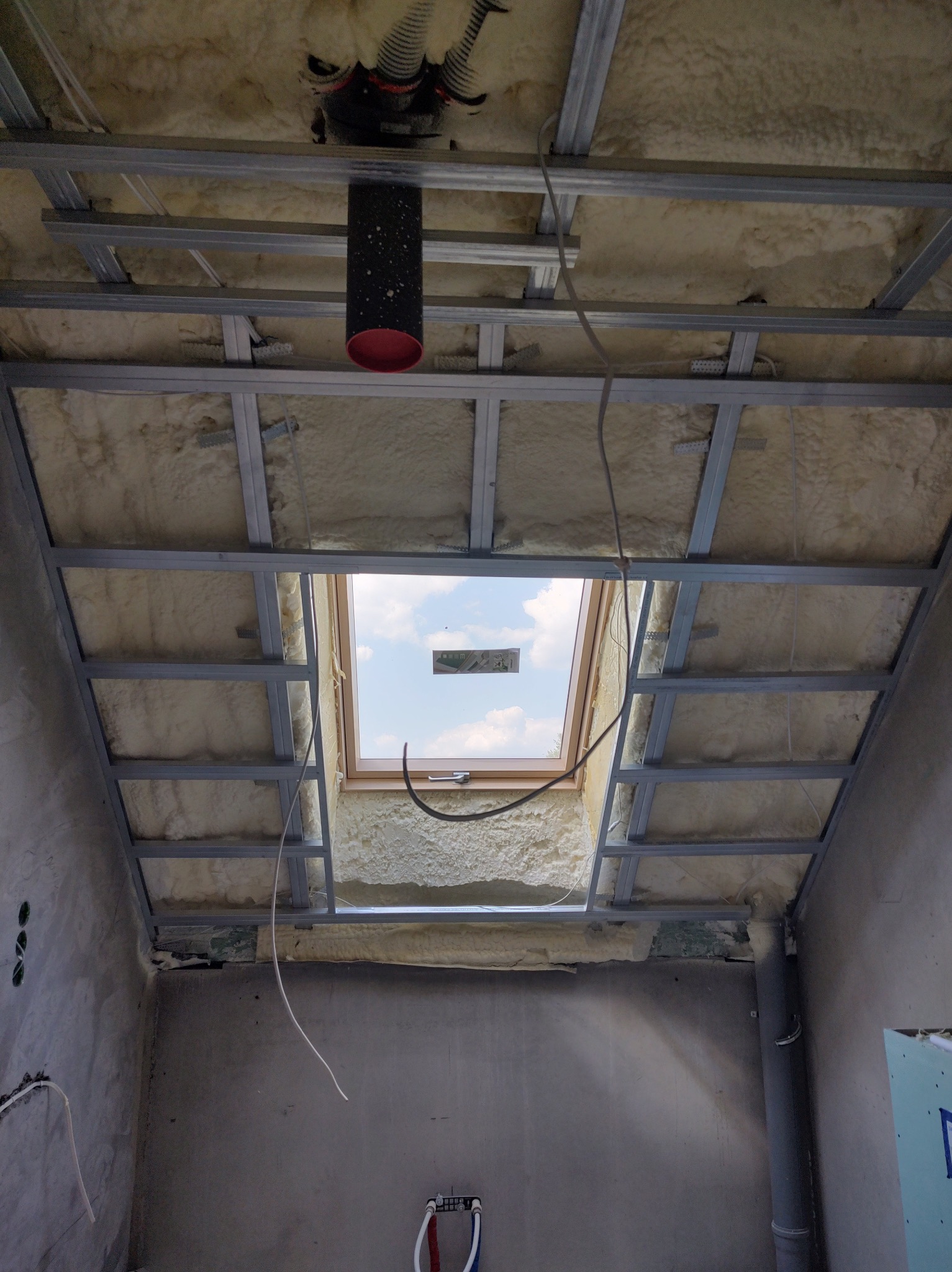 Poddasze w trakcie ocieplania pianką, widoczna konstrukcja metalowa sufitu podwieszanego i okno dachowe z widokiem na niebo.