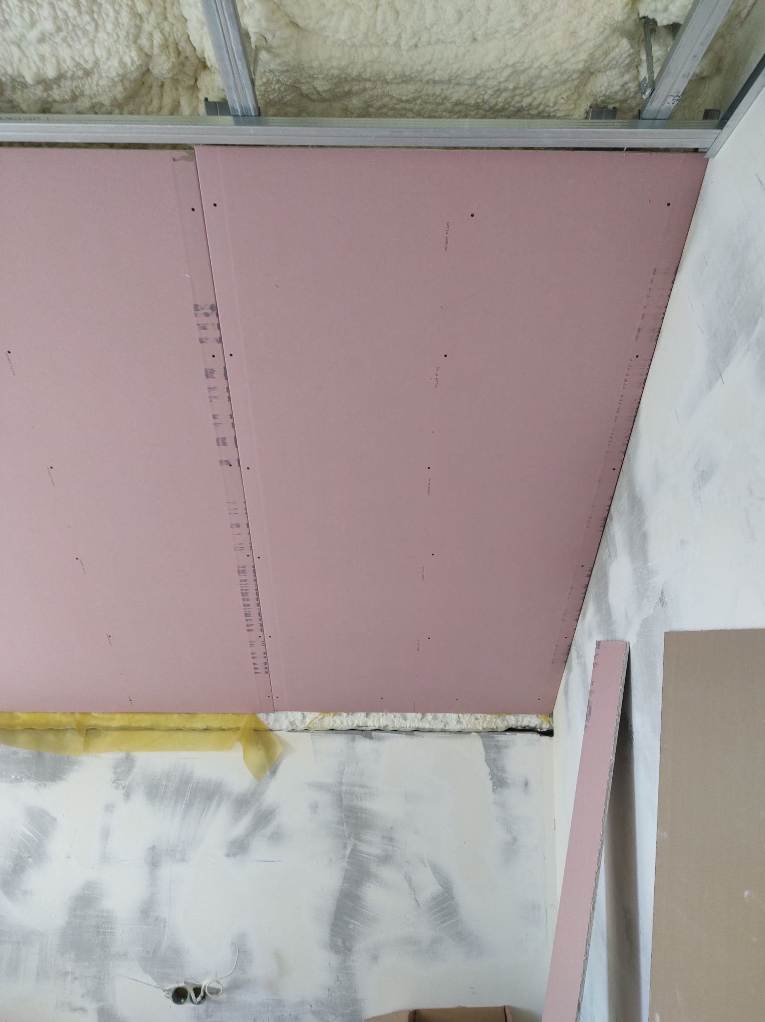 Montaż różowej płyty gipsowo-kartonowej na metalowym stelażu sufitowym, widoczne ocieplenie pianką między płytą a stropem, ściany w trakcie szpachlowania.