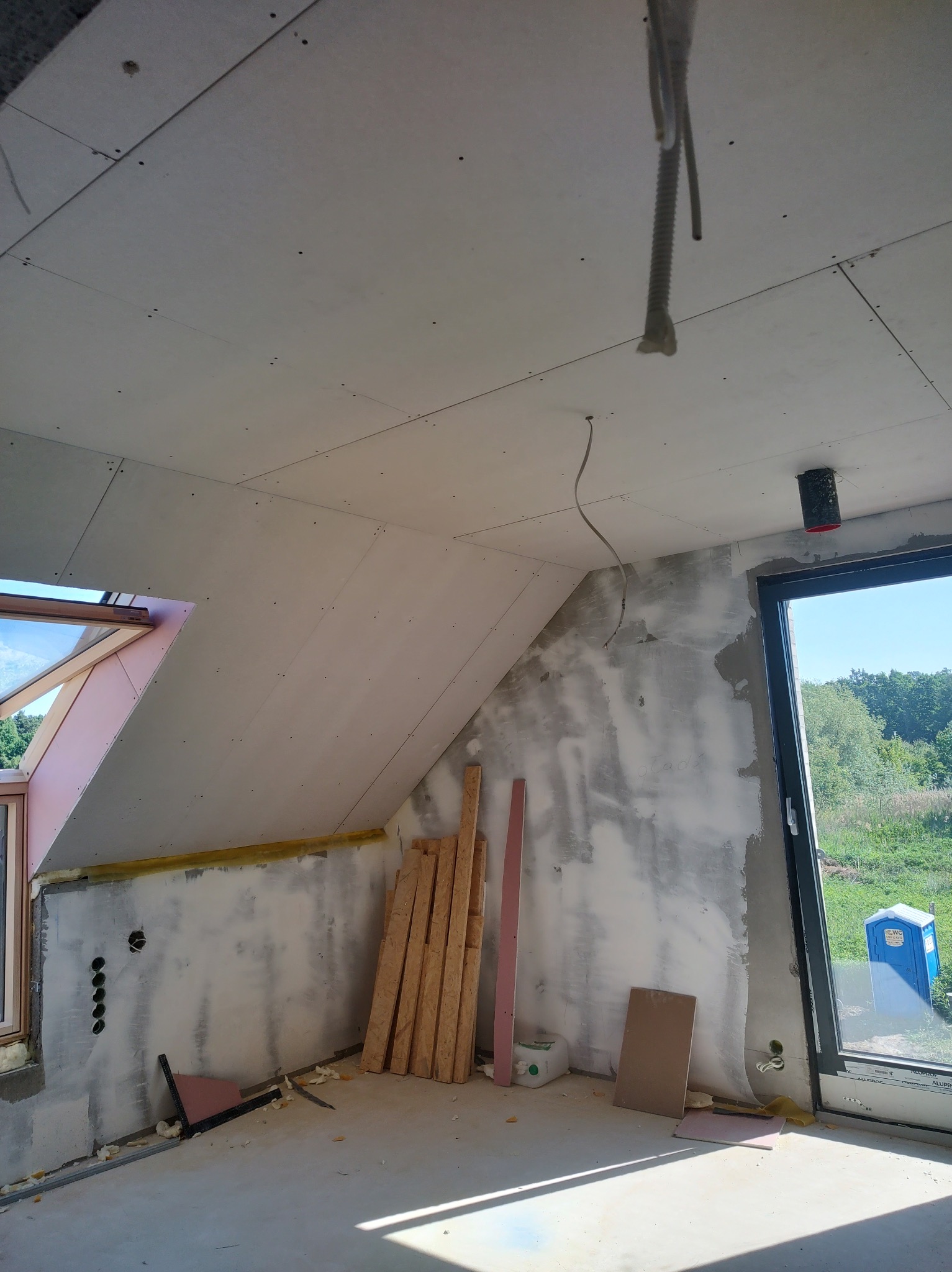 Poddasze w trakcie prac wykończeniowych: widoczne płyty kartonowo-gipsowe na skosach, przygotowane drewniane elementy konstrukcyjne, częściowo zagruntowane ściany oraz okno dachowe z widokiem...