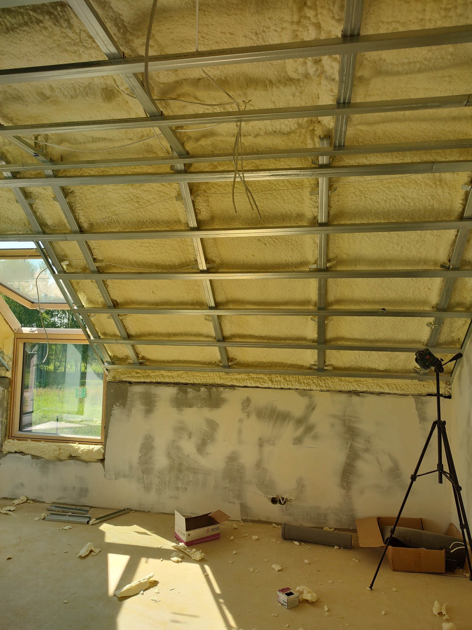 Poddasze w trakcie ocieplania pianą poliuretanową, widoczna konstrukcja metalowa sufitu, okno dachowe, ślady po pracach na ścianach, laser krzyżowy na statywie, resztki piany na podłodze.