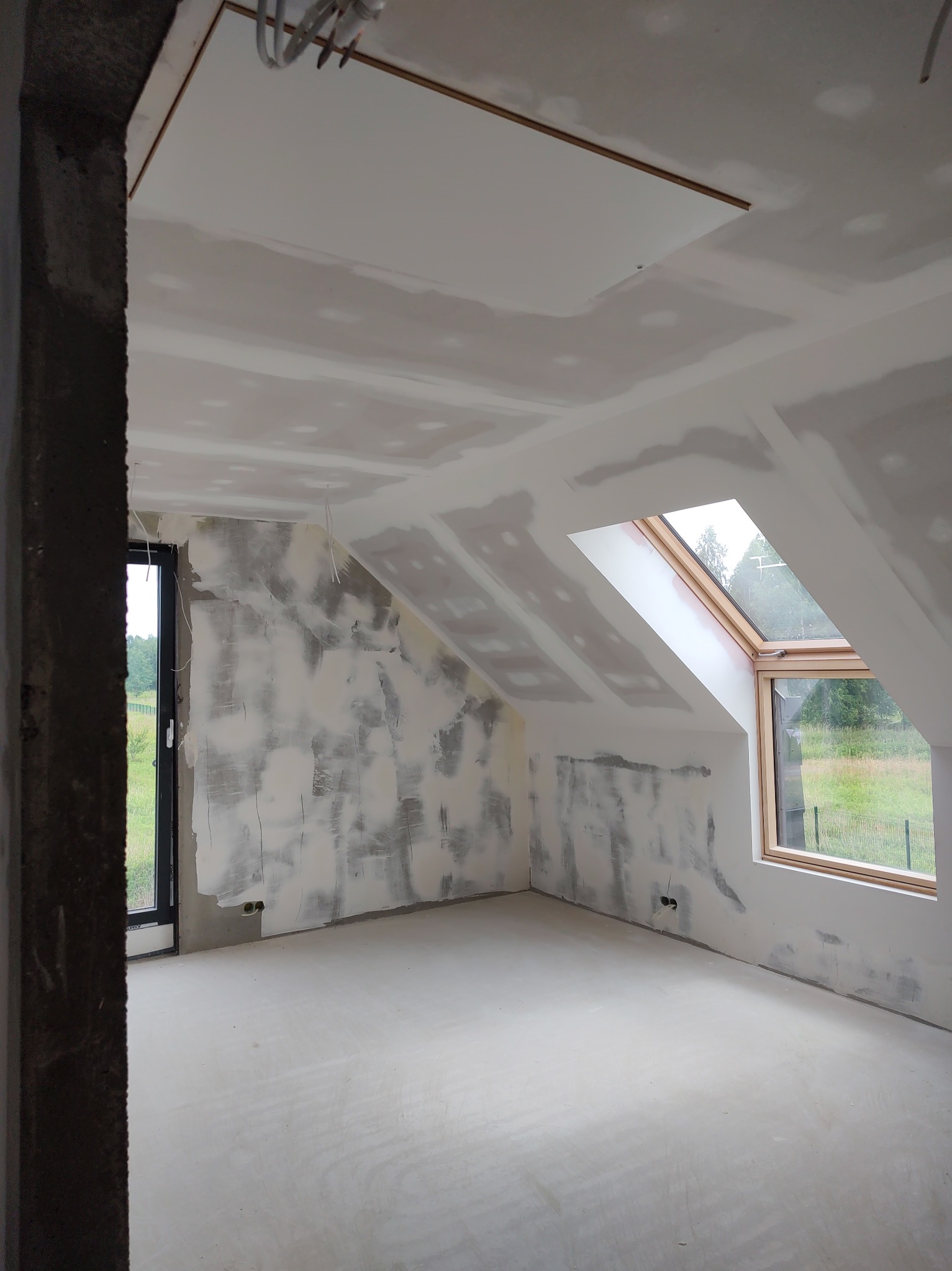 Poddasze w trakcie prac wykończeniowych: ściany z nałożoną warstwą gładzi, częściowo pokryte płytami kartonowo-gipsowymi, widoczne okno dachowe i surowy betonowy filar.