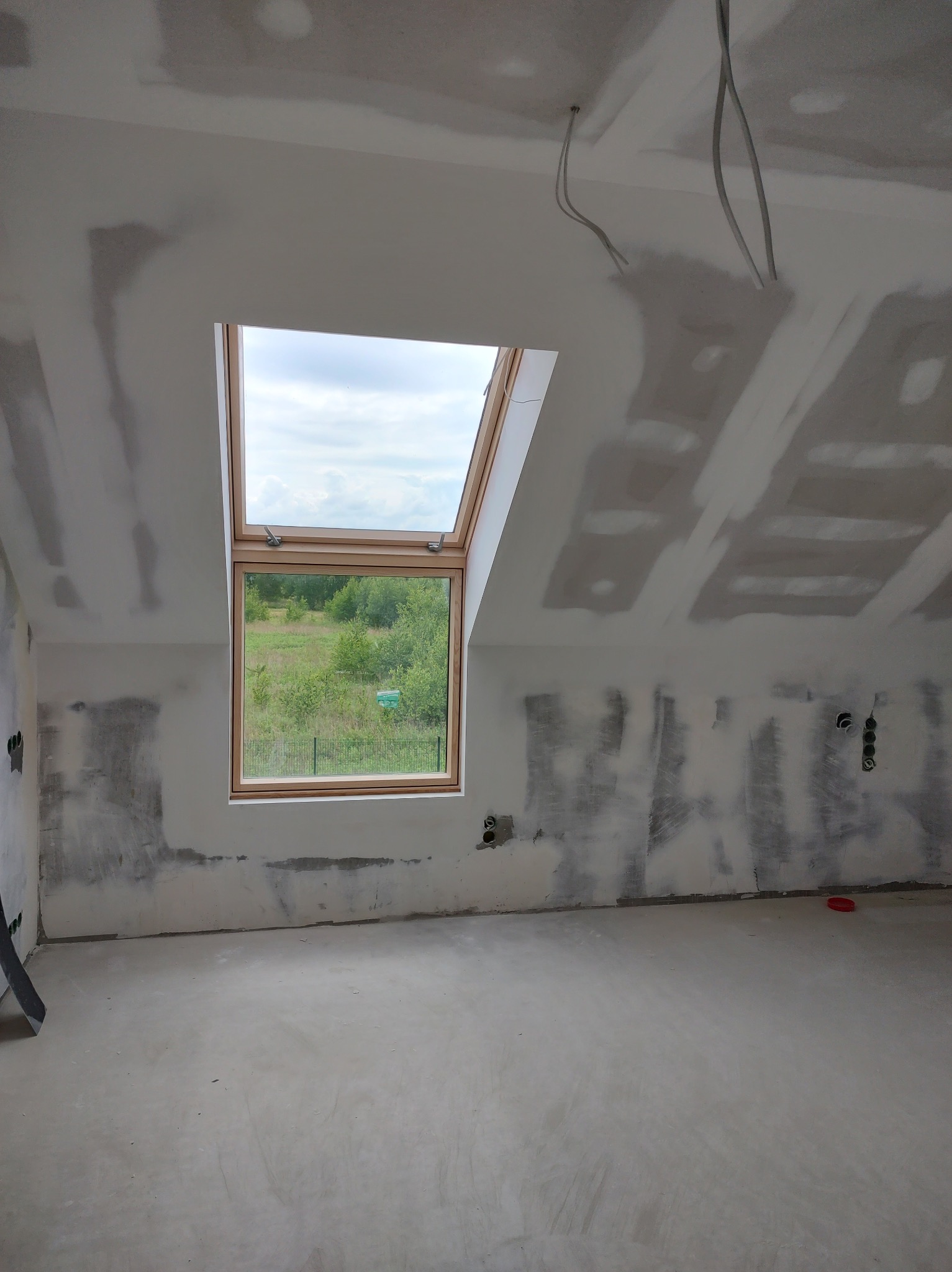 Poddasze w trakcie prac wykończeniowych, widoczne płyty kartonowo-gipsowe na skosach ścian i suficie, surowe gładzie, okno dachowe z widokiem na zieleń, wystające kable elektryczne.
