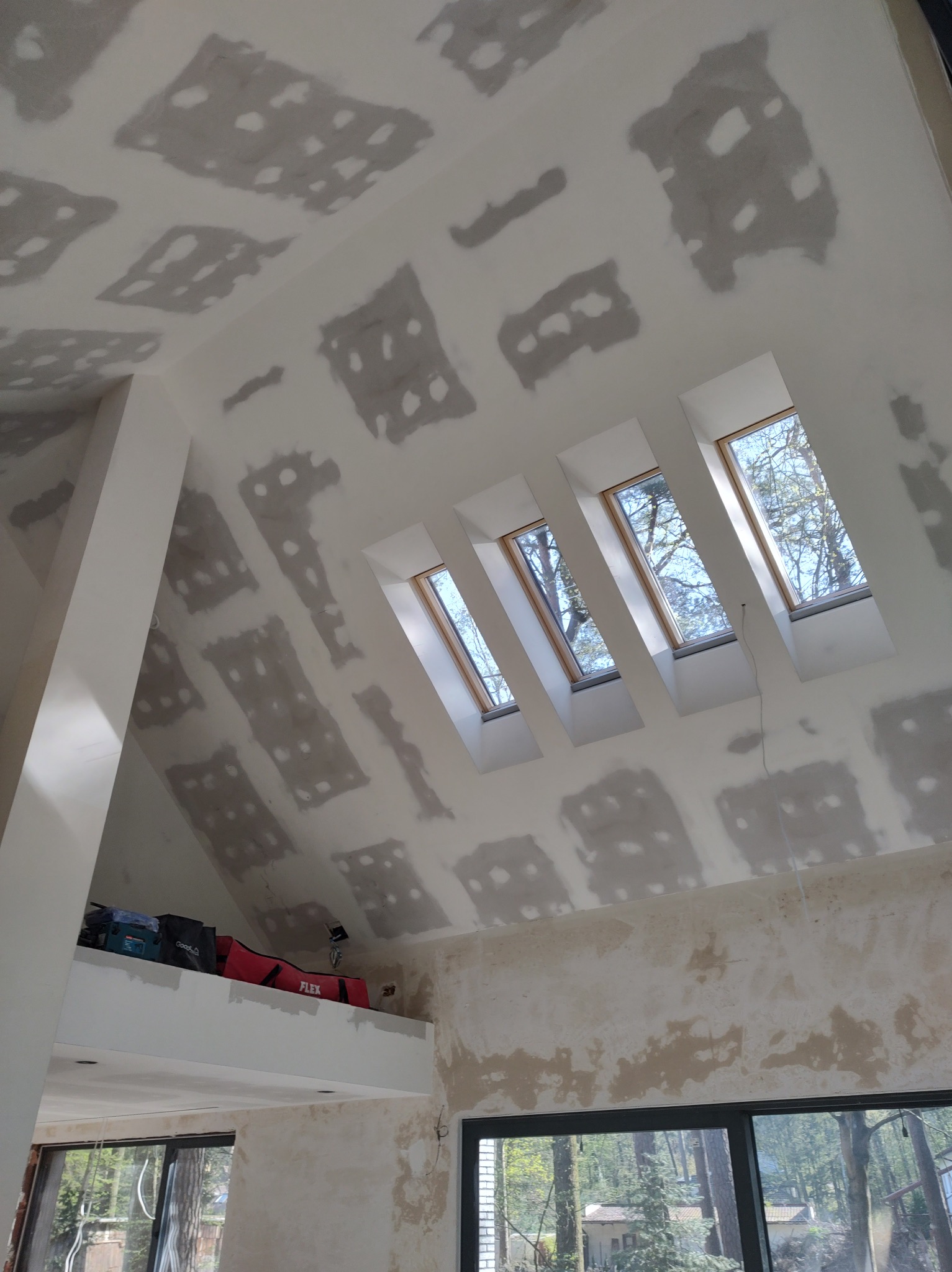 Poddasze w trakcie przygotowania ścian i sufitu do dalszych prac wykończeniowych, widoczne łaty szpachli na płytach gipsowo-kartonowych, cztery okna dachowe w rzędzie oraz narzędzia na antresoli.