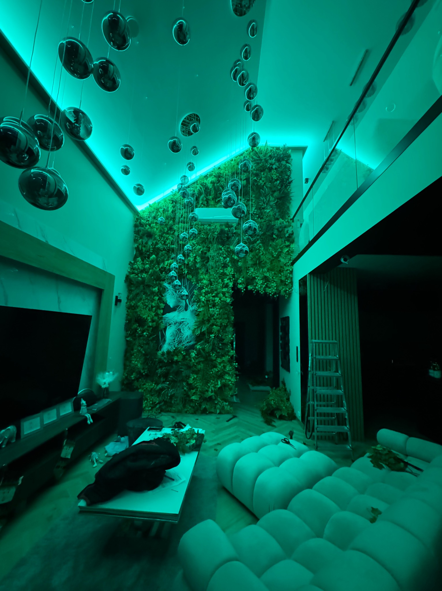 Nowoczesne wnętrze z pionową ścianą zieleni, oświetleniem LED i wiszącymi dekoracjami. Widoczna sofa i drabina, sugerująca prace wykończeniowe.