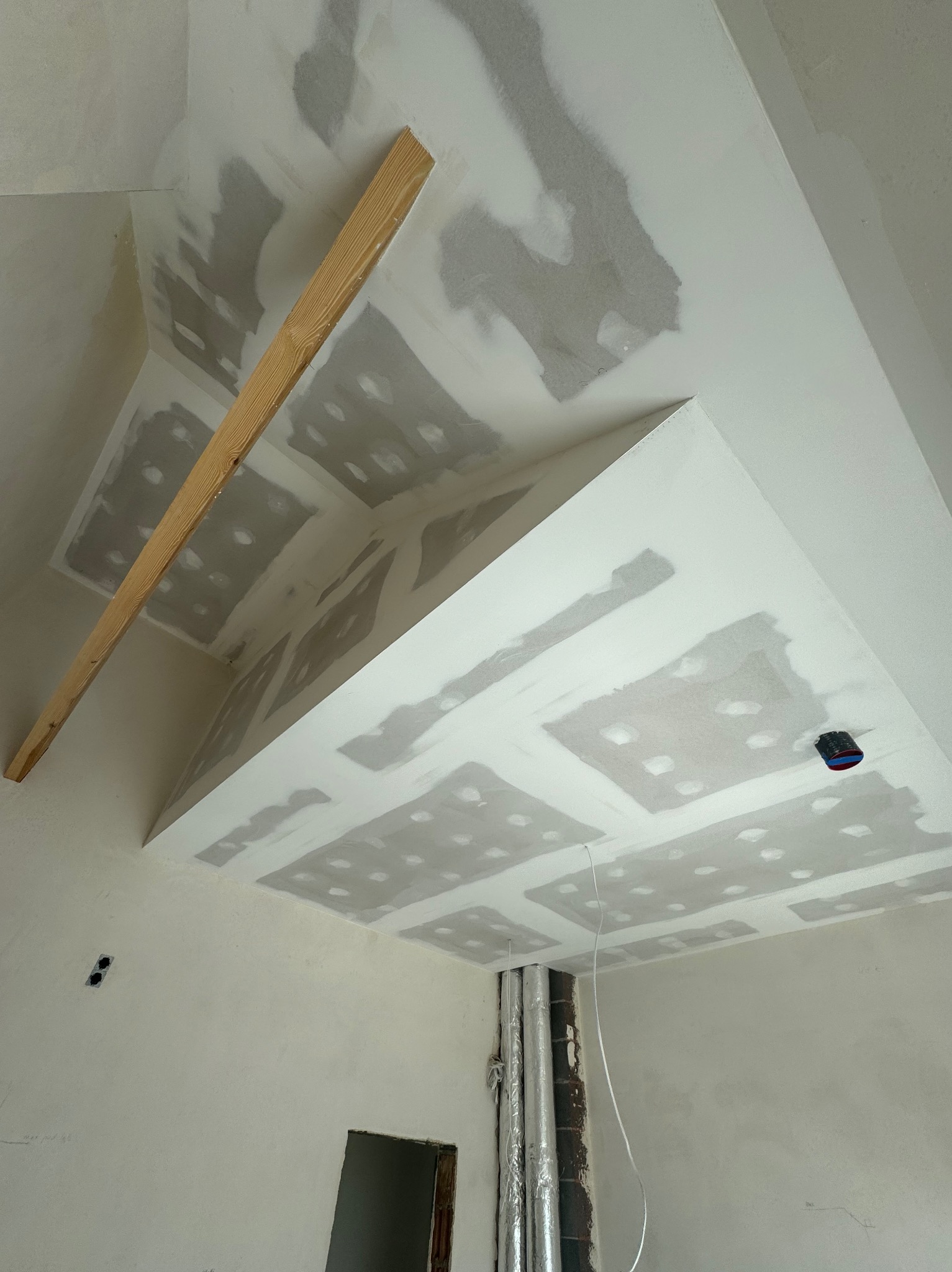 Sufit podwieszany z płyt karton-gips na etapie szpachlowania, widoczne łączenia płyt. Fragment drewnianej listwy na suficie. Instalacja elektryczna i rury w tle.