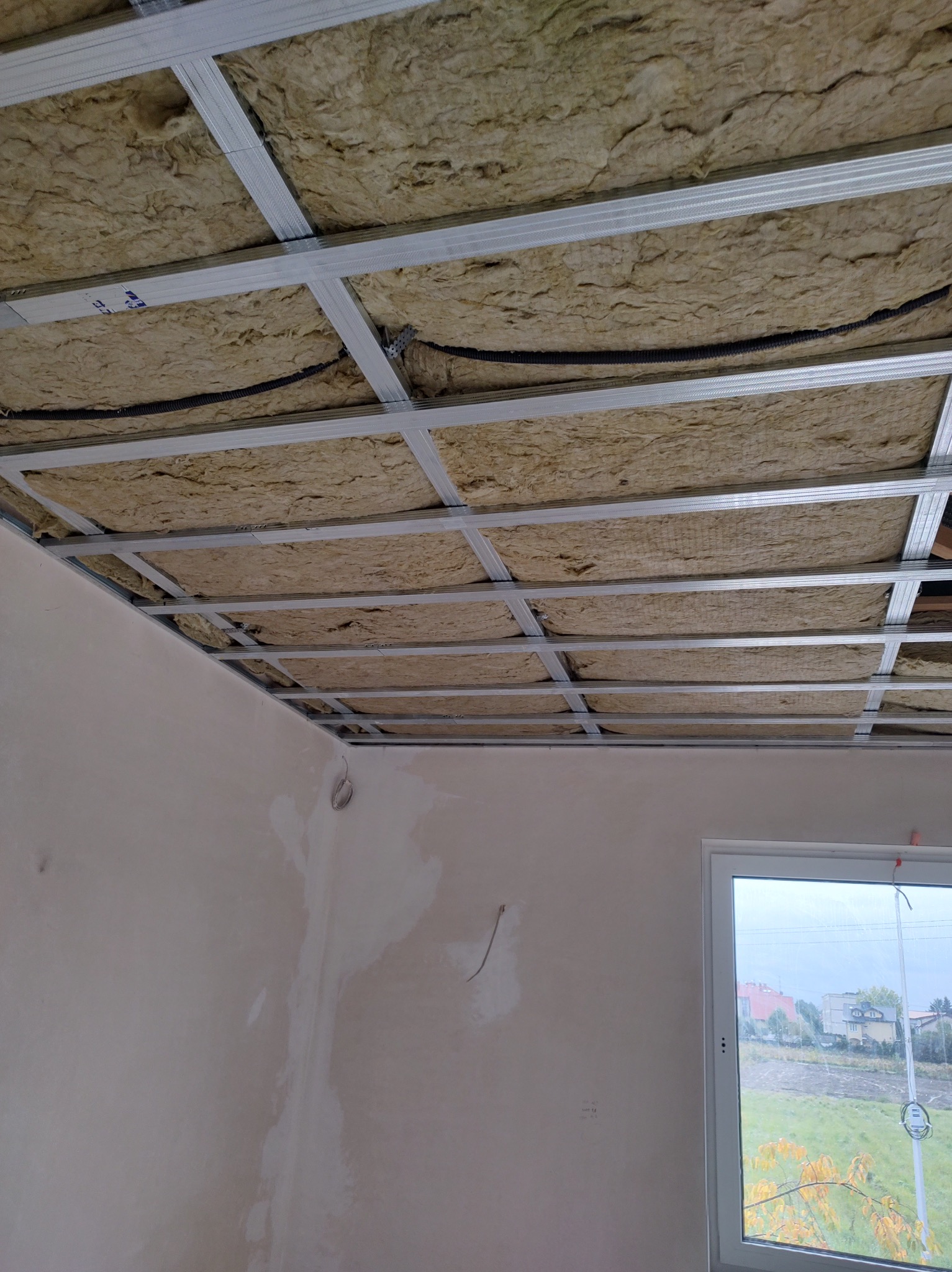 Montaż izolacji z wełny mineralnej na suficie podwieszanym, widoczne profile metalowe i przewody elektryczne. Ściana w surowym stanie, okno z widokiem na zewnątrz.