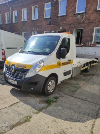 Biała laweta Renault Master z żółtymi pasami i napisem 'Pomoc Drogowa' zaparkowana na betonowym placu przed ceglanym budynkiem mieszkalnym.