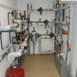 Złożona instalacja rur i zaworów z manometrami w pomieszczeniu technicznym, widoczny czerwony zbiornik wyrównawczy i odpływ podłogowy