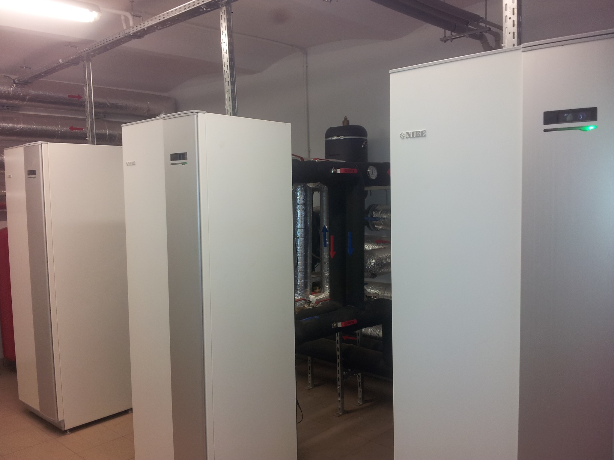 Trzy białe pompy ciepła Nibe w kotłowni, widoczne rury instalacji grzewczej z izolacją i zaworami, oraz metalowe koryta kablowe pod sufitem.