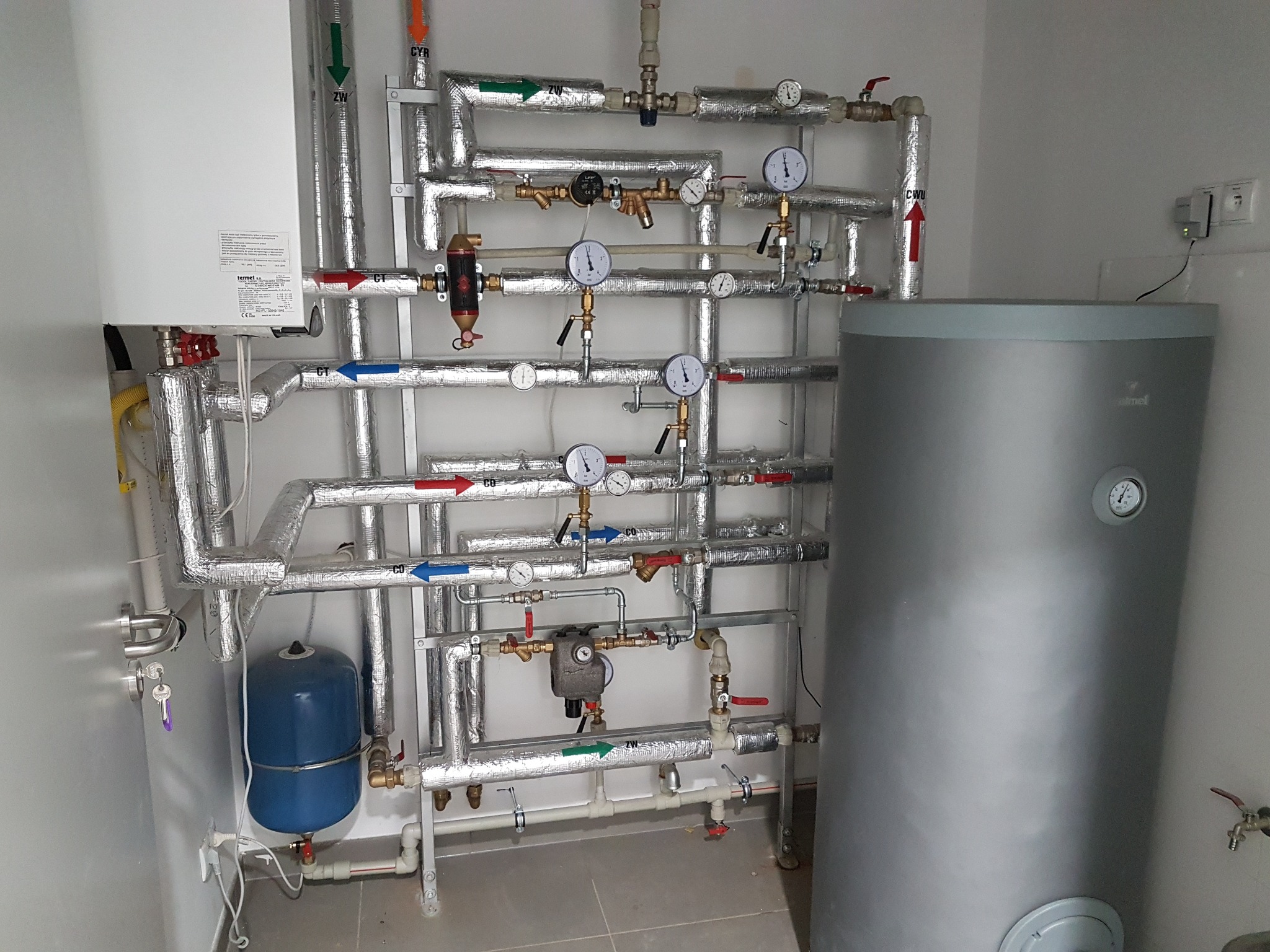 Węzeł cieplny w kotłowni: srebrny zbiornik akumulacyjny, biały piec, niebieskie naczynie przeponowe i rozbudowana instalacja rur w izolacji aluminiowej z manometrami i zaworami.