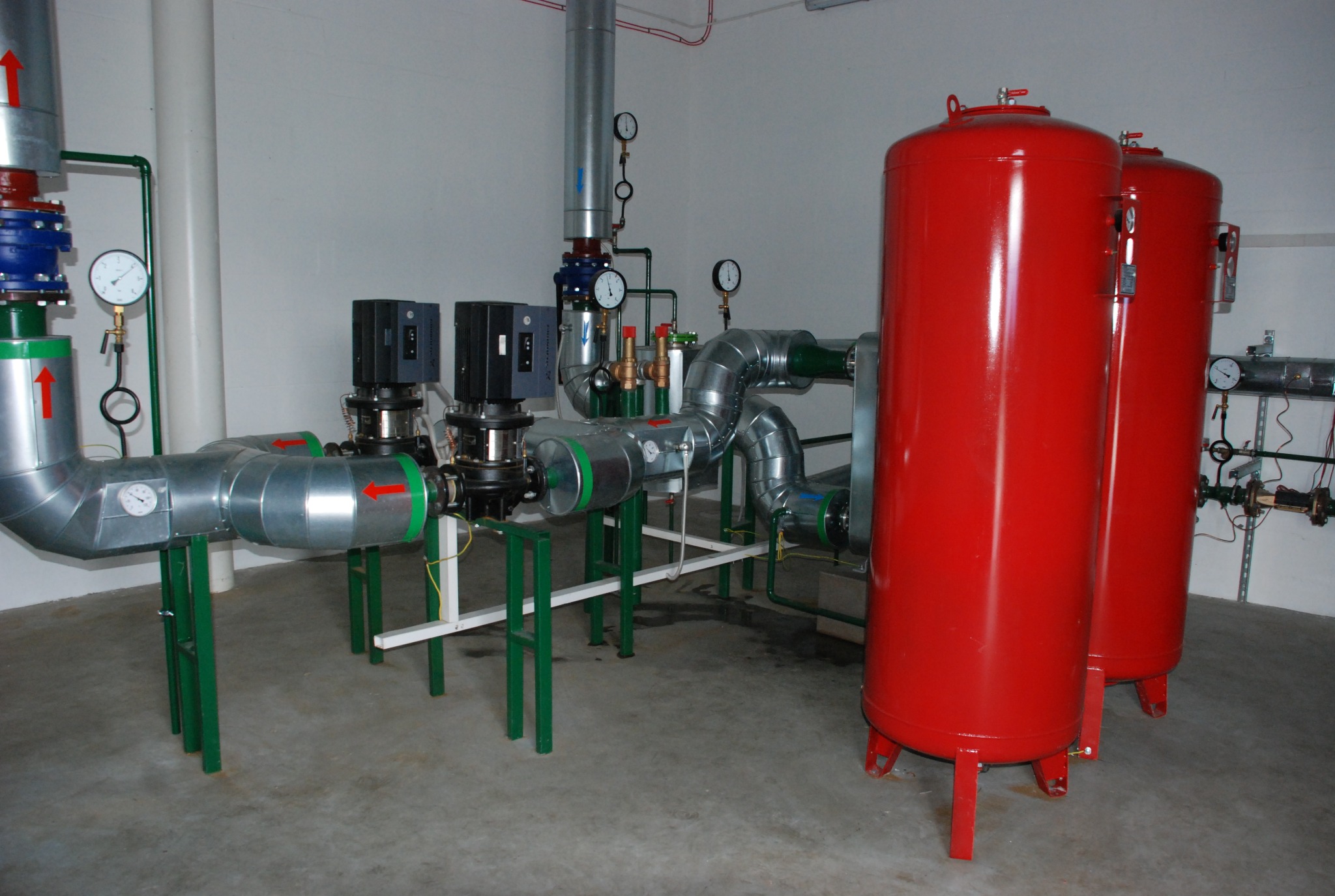 Dwa pionowe, czerwone zbiorniki ciśnieniowe, połączone systemem rur z manometrami, zainstalowane w pomieszczeniu z betonową podłogą i białymi ścianami.