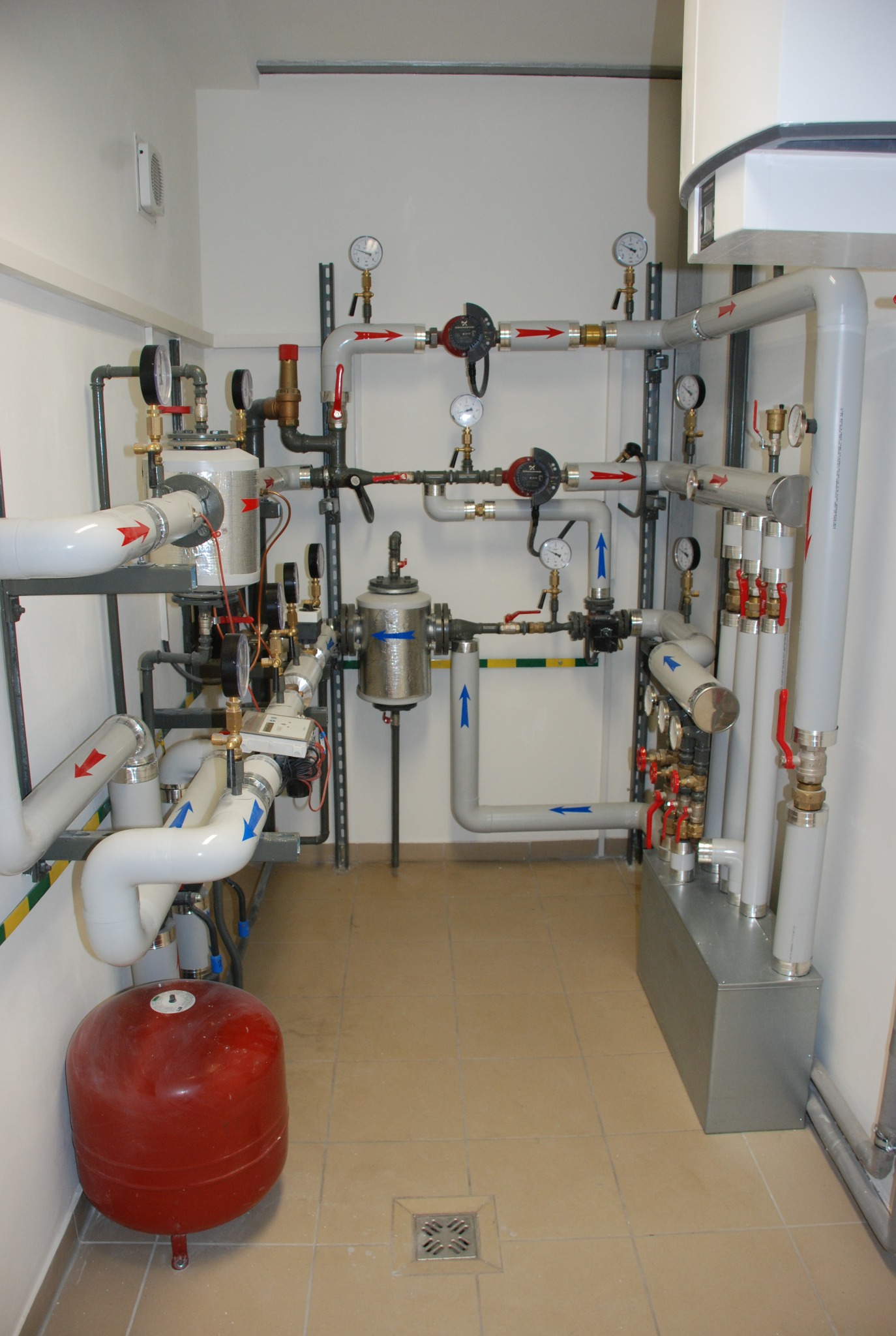 Złożona instalacja rur i zaworów z manometrami w pomieszczeniu technicznym, widoczny czerwony zbiornik wyrównawczy i odpływ podłogowy