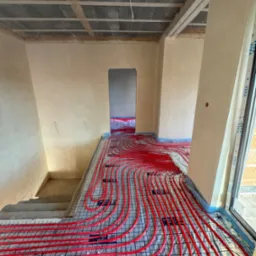 Czerwone rury ogrzewania podłogowego ułożone na siatce w surowym wnętrzu domu w trakcie budowy, widoczne schody i okno.