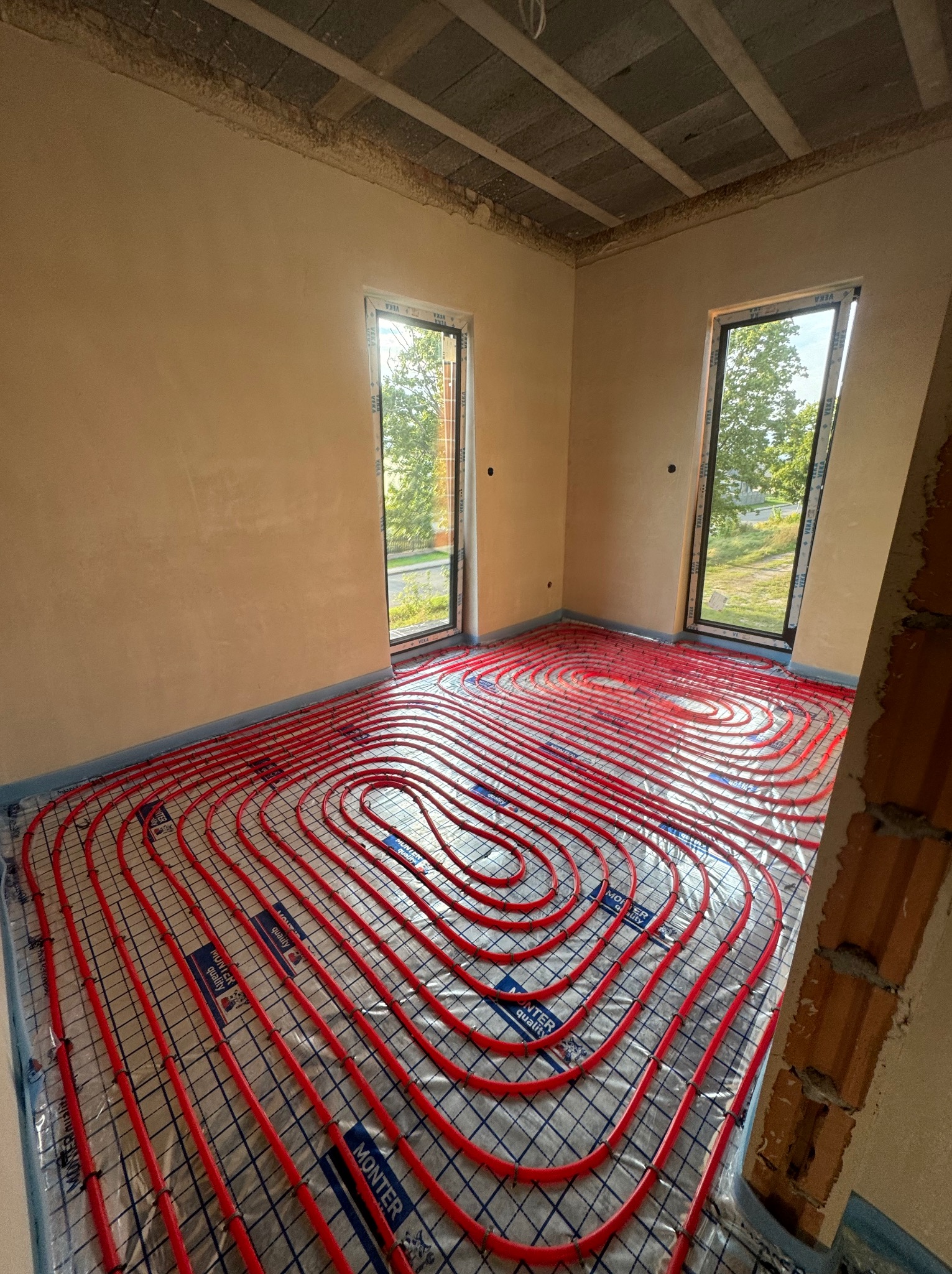 Czerwone rury ogrzewania podłogowego ułożone spiralnie na folii w surowym pomieszczeniu z dwoma oknami, widoczne niedokończone ściany i surowy strop.