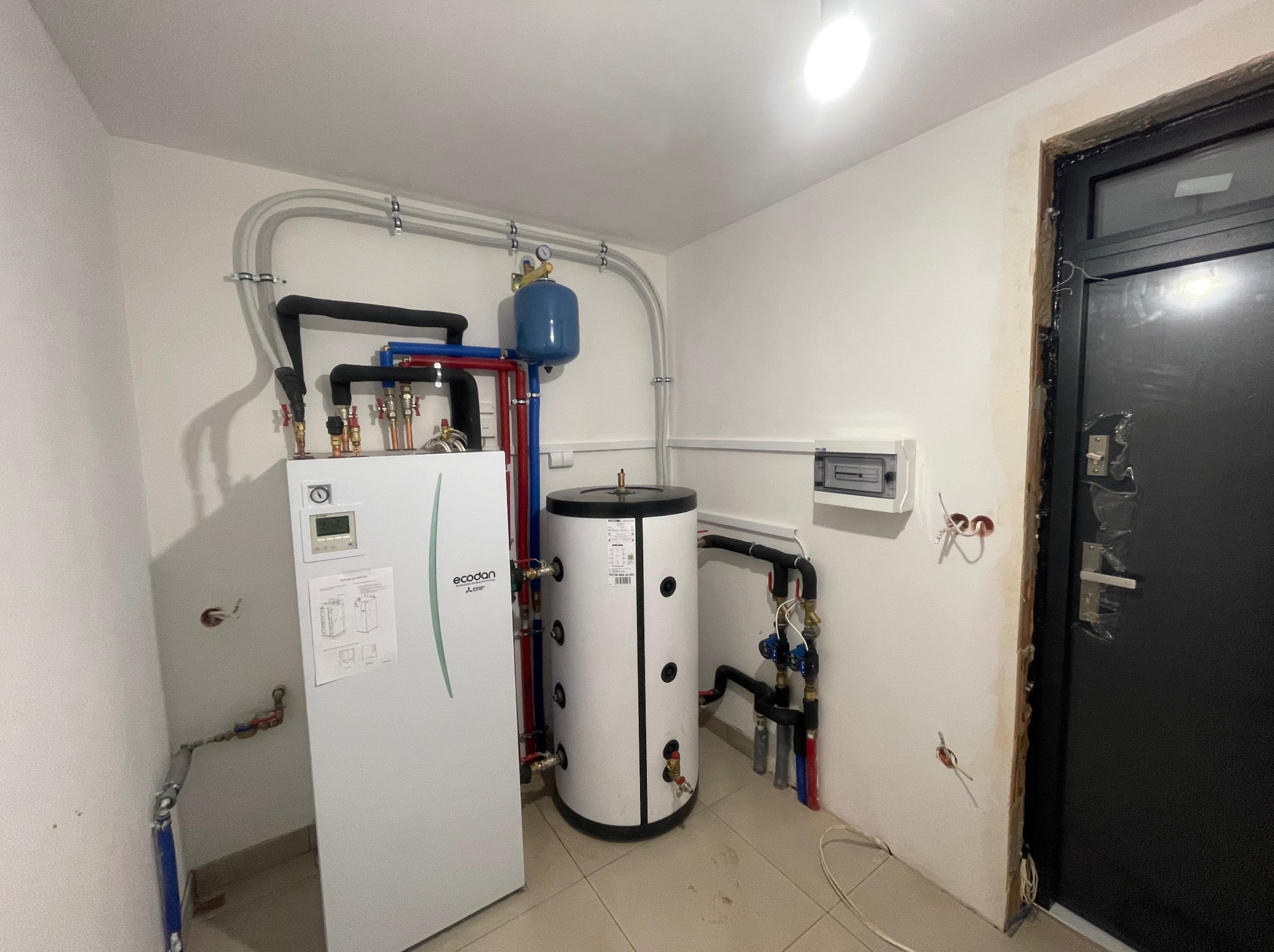Biała pompa ciepła Ecodan i zbiornik buforowy w kotłowni, widoczne rury w izolacji, manometr na zbiorniku wyrównawczym, instalacja elektryczna w peszlach, surowa ściana przy futrynie drzwi.