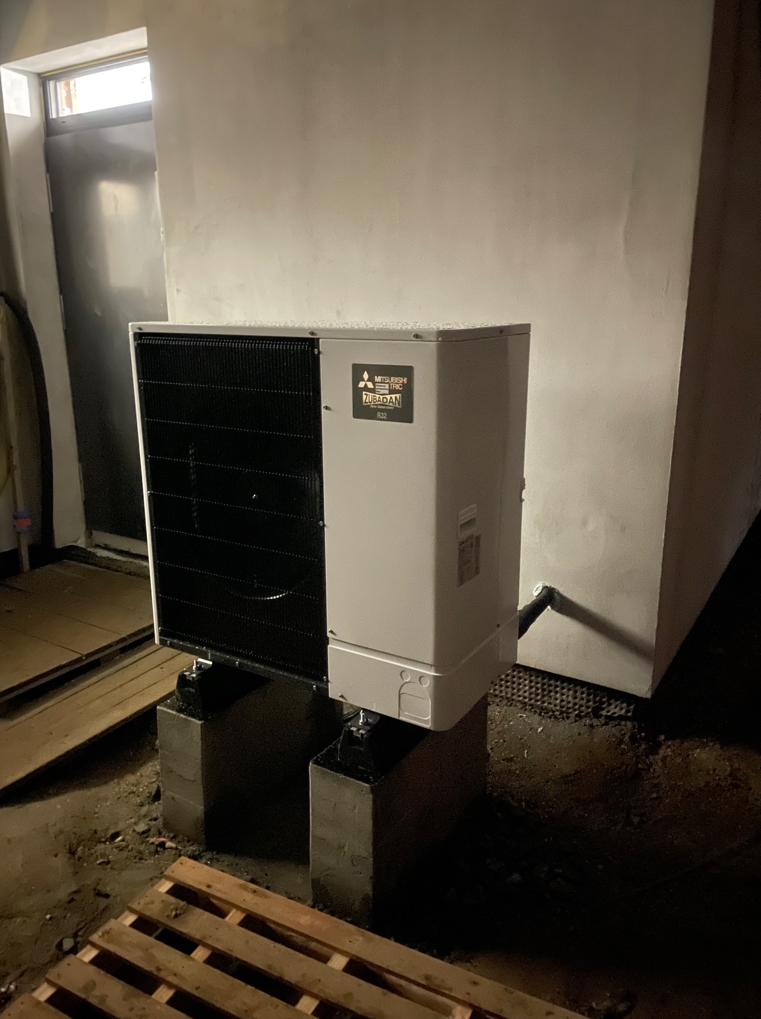 Zewnętrzna jednostka pompy ciepła Mitsubishi Zubadan ustawiona na betonowych bloczkach w surowym pomieszczeniu, widoczne rury i paleta na podłodze.