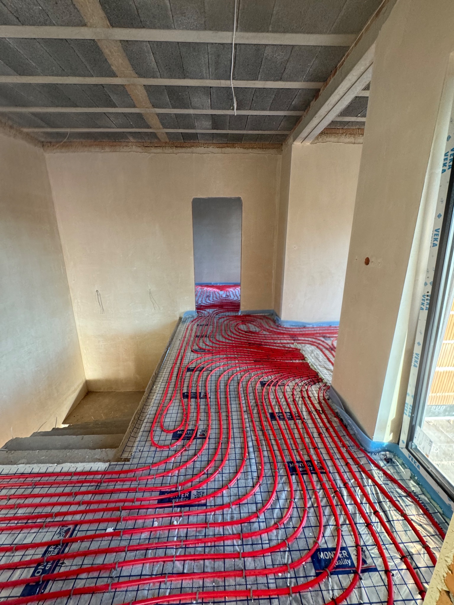 Czerwone rury ogrzewania podłogowego ułożone na siatce w surowym wnętrzu domu w trakcie budowy, widoczne schody i okno.
