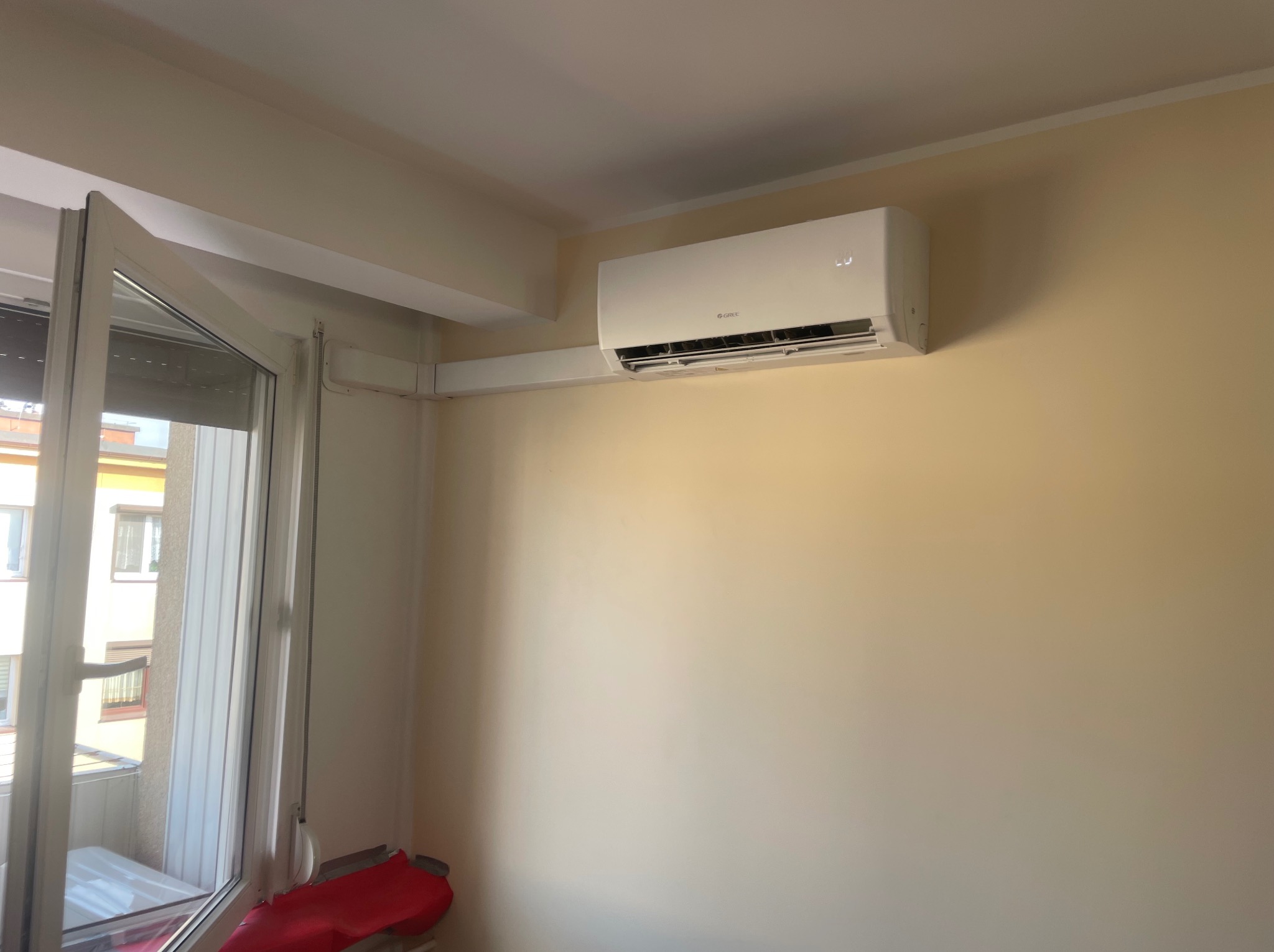 Biały klimatyzator marki Gree zamontowany na beżowej ścianie w pokoju z otwartym oknem balkonowym.