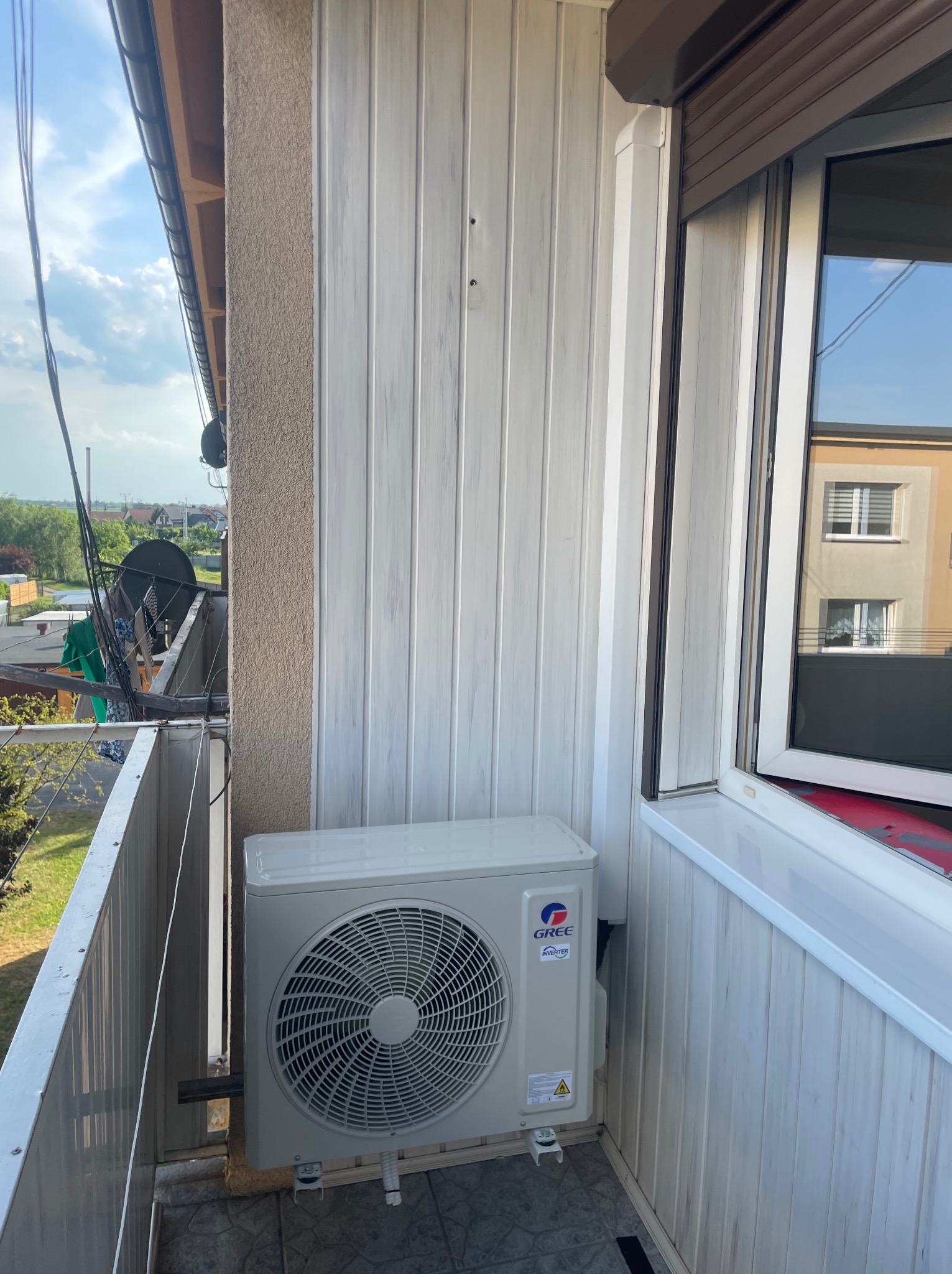 Zewnętrzna jednostka klimatyzacji marki Gree zamontowana na balkonie, widok na okoliczne budynki i zieleń.
