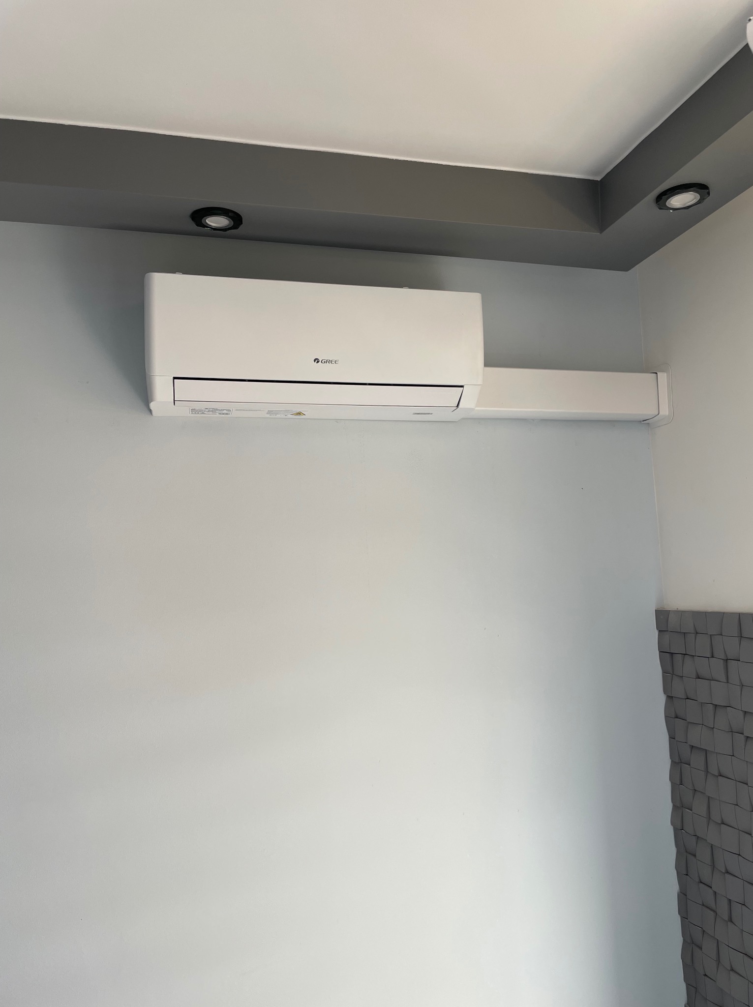 Biały klimatyzator marki Gree zamontowany na jasnoszarej ścianie, z widoczną maskownicą instalacji i fragmentem dekoracyjnej ściany z trójwymiarowych paneli w dolnym prawym rogu.
