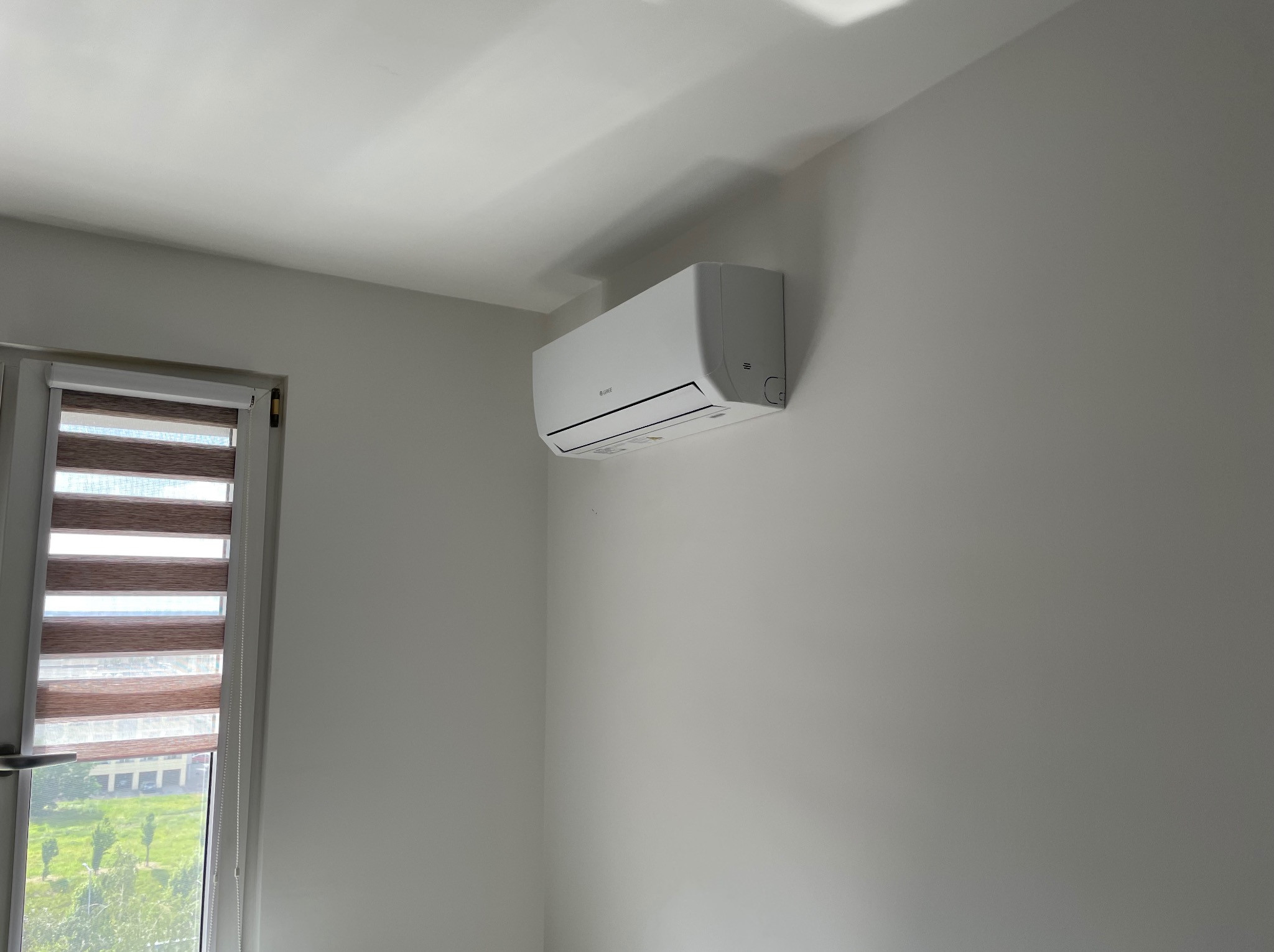 Biała jednostka klimatyzacyjna marki Gree zamontowana na jasnej ścianie w pokoju z oknem z roletami dzień-noc.