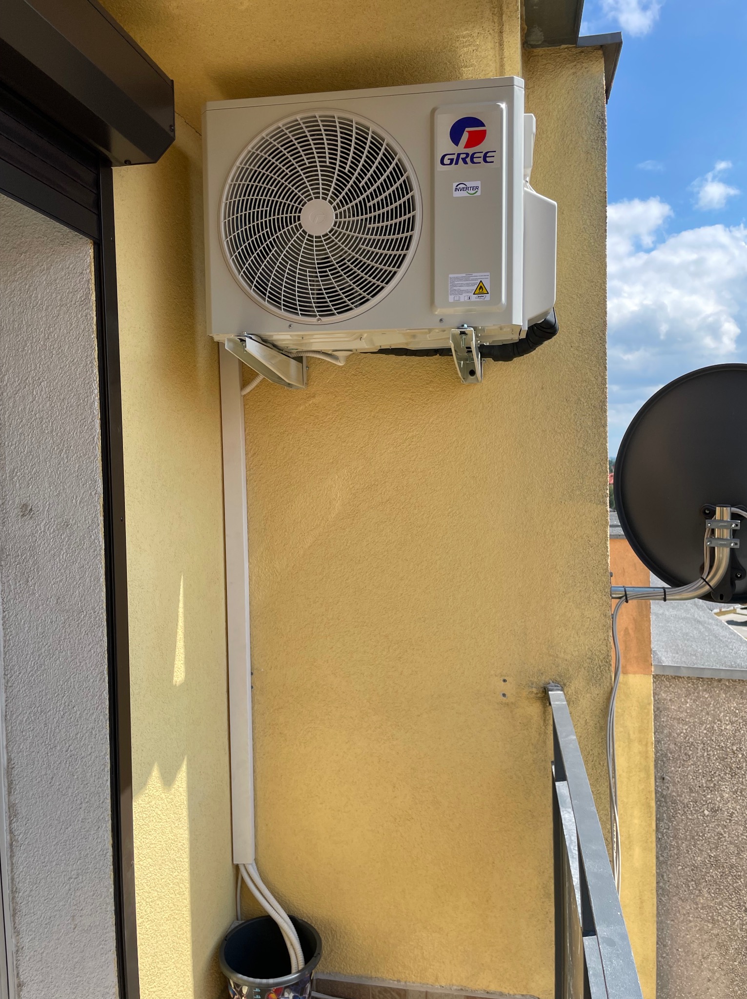 Zewnętrzna jednostka klimatyzacji GREE zamontowana na żółtej ścianie balkonu, widoczne białe korytko instalacyjne i wiadro z rurami odprowadzającymi skropliny.
