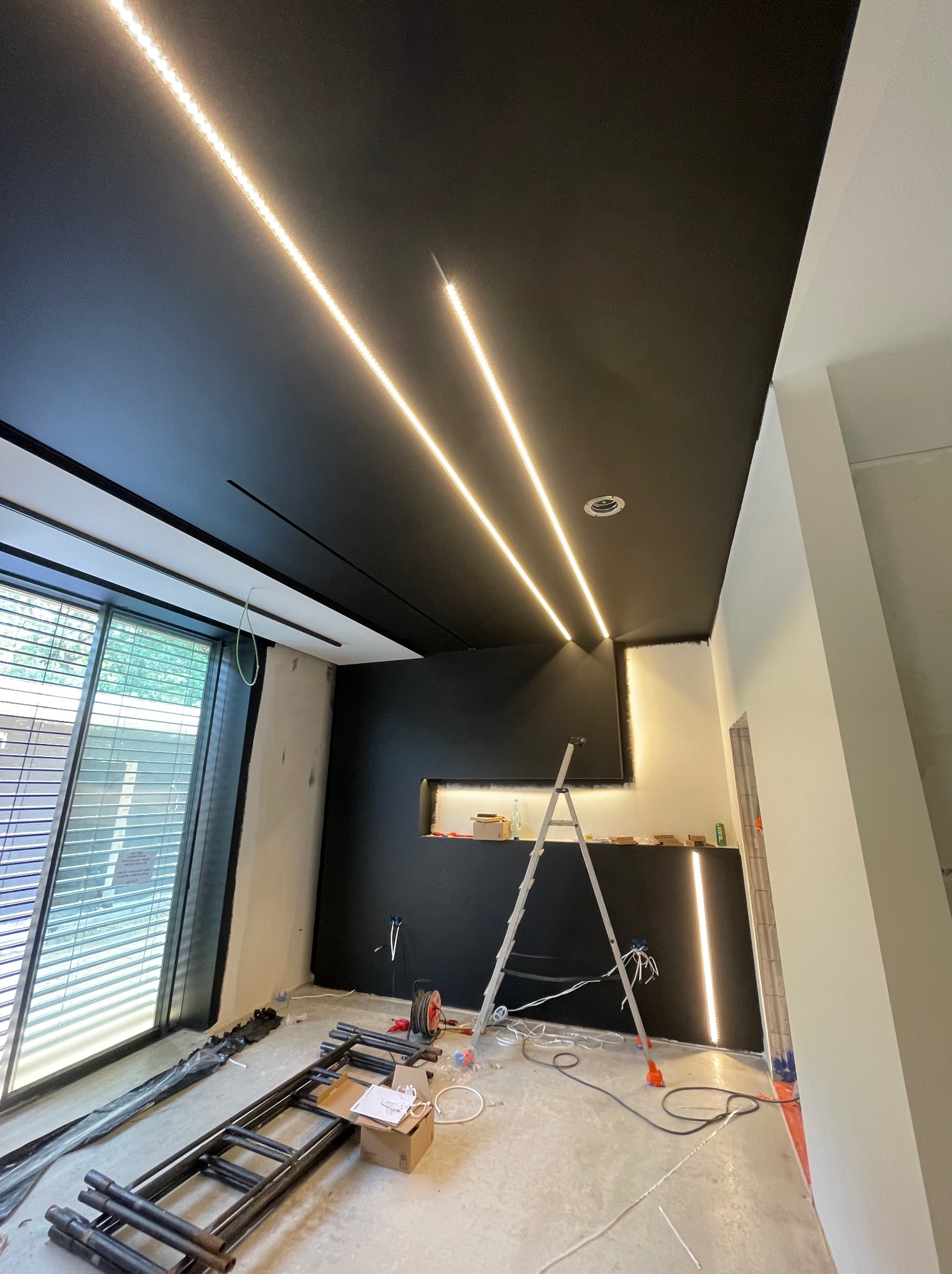 Wnętrze w trakcie wykańczania: czarny sufit z dwiema liniami oświetlenia LED, drabina oparta o ścianę, widoczne przewody i narzędzia, okno z żaluzjami.