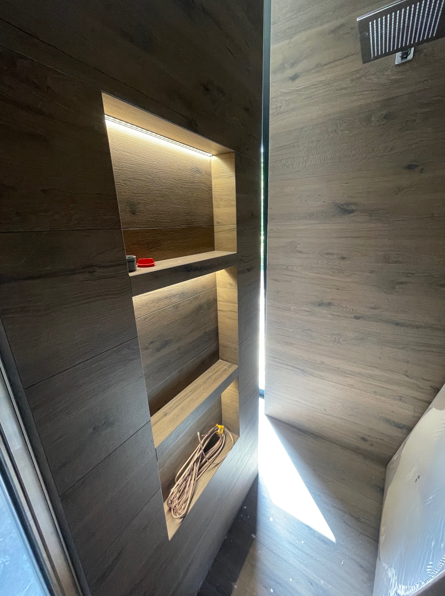 Nowoczesna łazienka z drewnopodobnymi płytkami, wnękami z podświetleniem LED i widoczną słuchawką prysznicową.