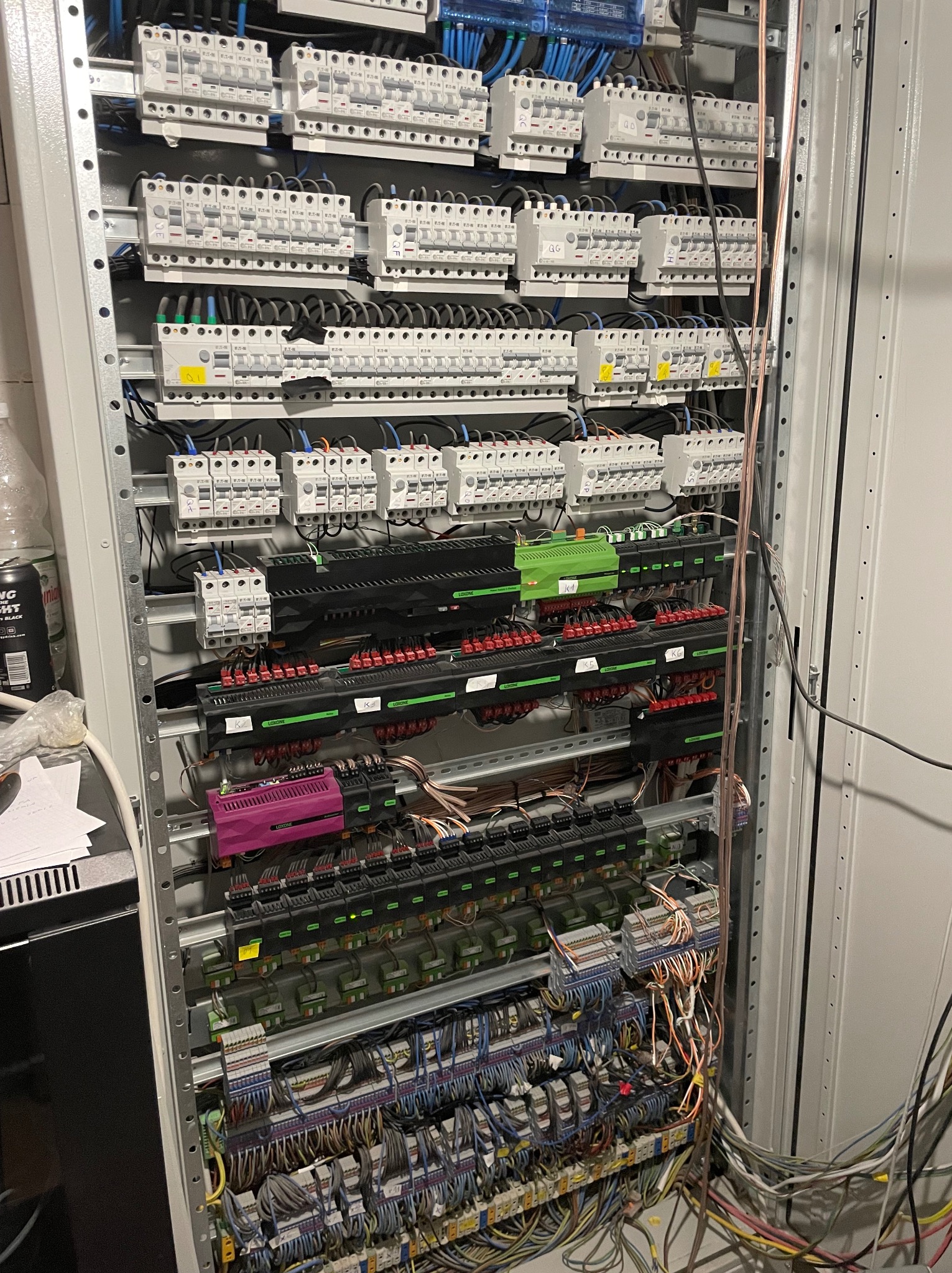 Rozdzielnica elektryczna z licznymi bezpiecznikami, modułami sterującymi Loxone (K1, K2, K5, K6) i gęstą siecią przewodów wewnątrz metalowej szafy.