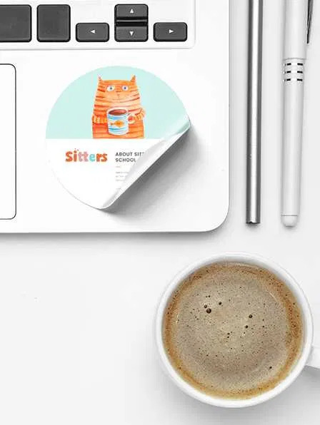 Naklejka z rysunkiem kota pijącego kawę na laptopie obok filiżanki kawy i długopisu.