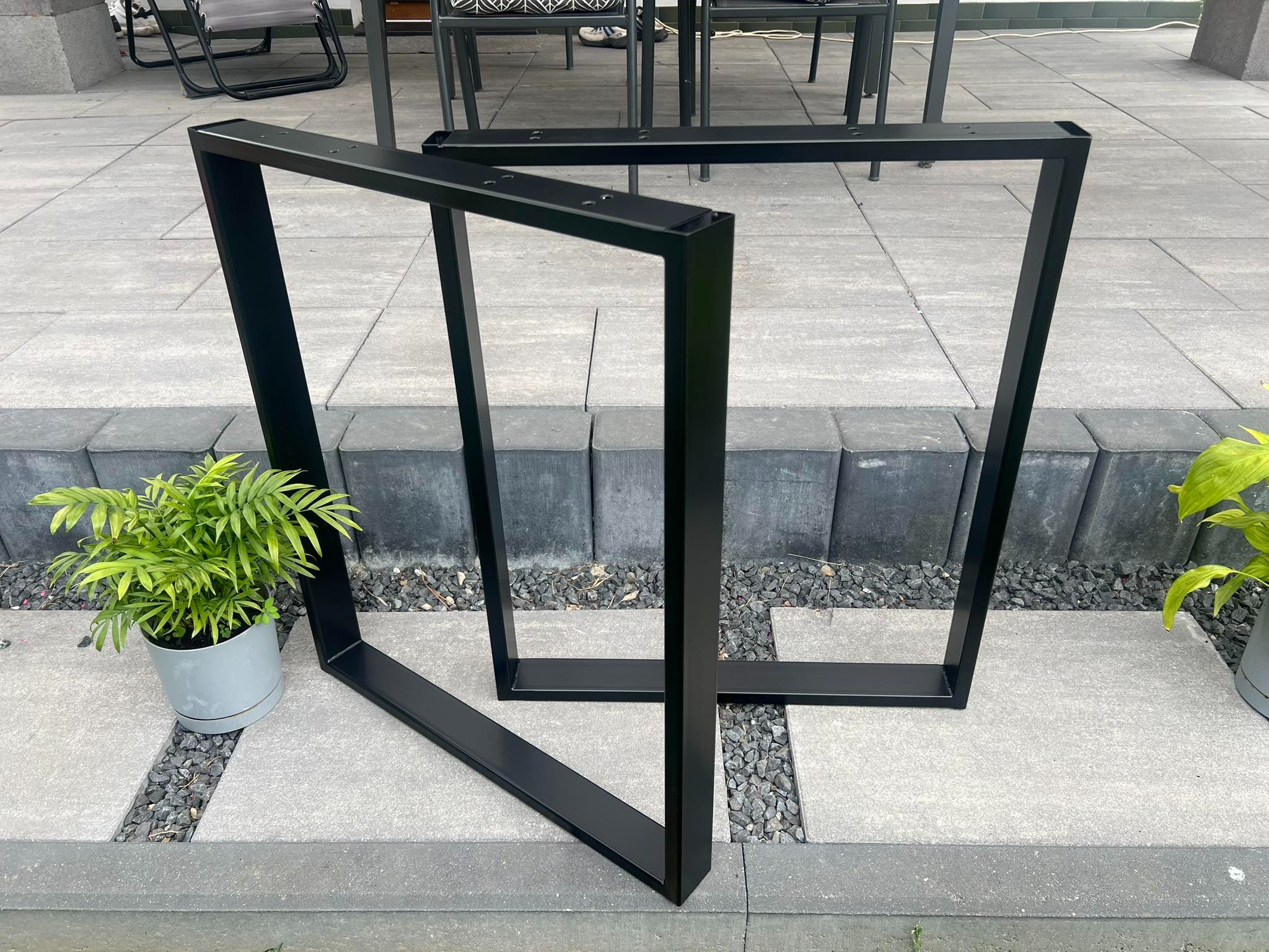 Dwie czarne, metalowe ramy trapezowe na tarasie z kostki, obok doniczka z zieloną rośliną. Elementy do stołu lub ławki ogrodowej.