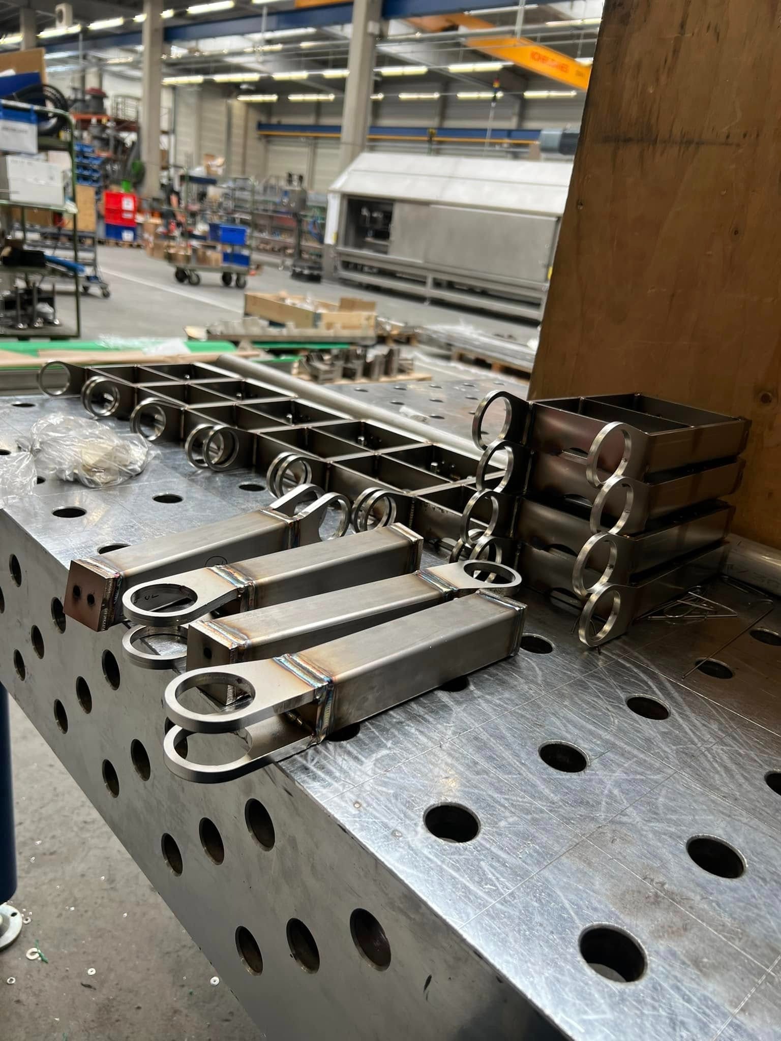 Metalowe elementy konstrukcyjne z widocznymi spawami, ułożone na perforowanym stole warsztatowym w hali produkcyjnej. Elementy o różnych kształtach, niektóre z okrągłymi otworami.