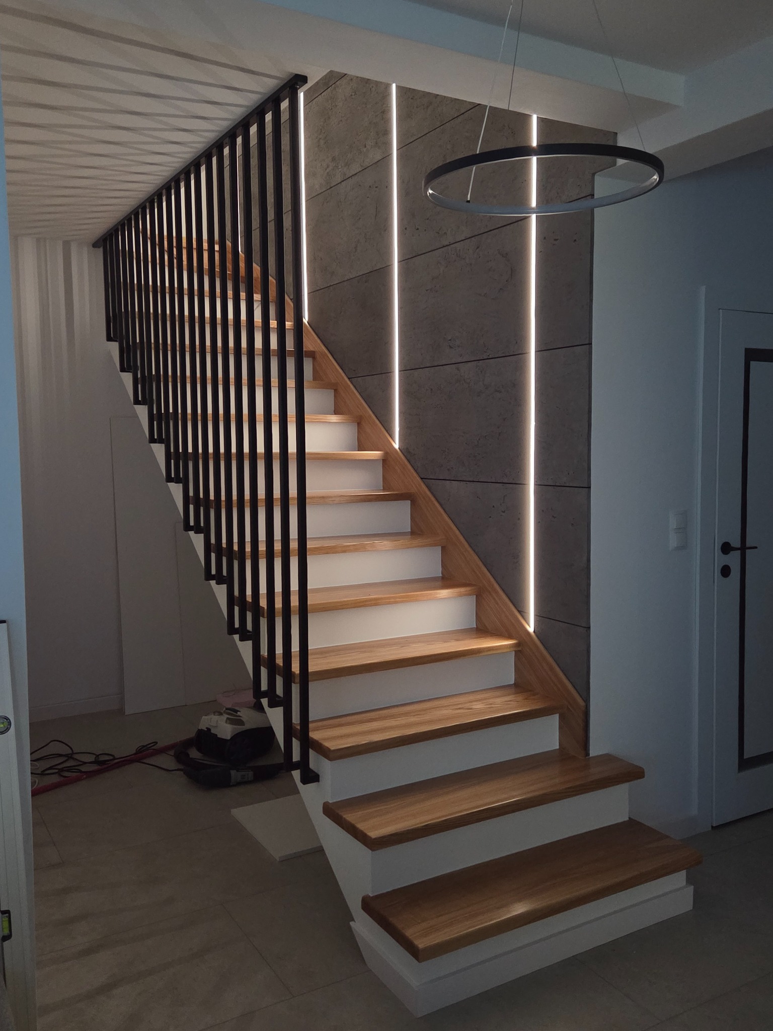 Nowoczesne schody drewniane z czarną, minimalistyczną balustradą, oświetleniem LED w ścianie i okrągłą lampą sufitową. Widok z dołu schodów.