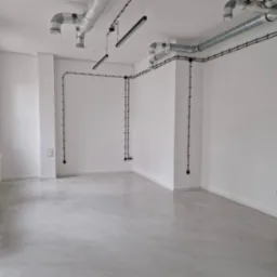 Jasnoszara, gładka posadzka w pustym, białym pomieszczeniu z widocznymi instalacjami elektrycznymi i wentylacyjnymi pod sufitem.