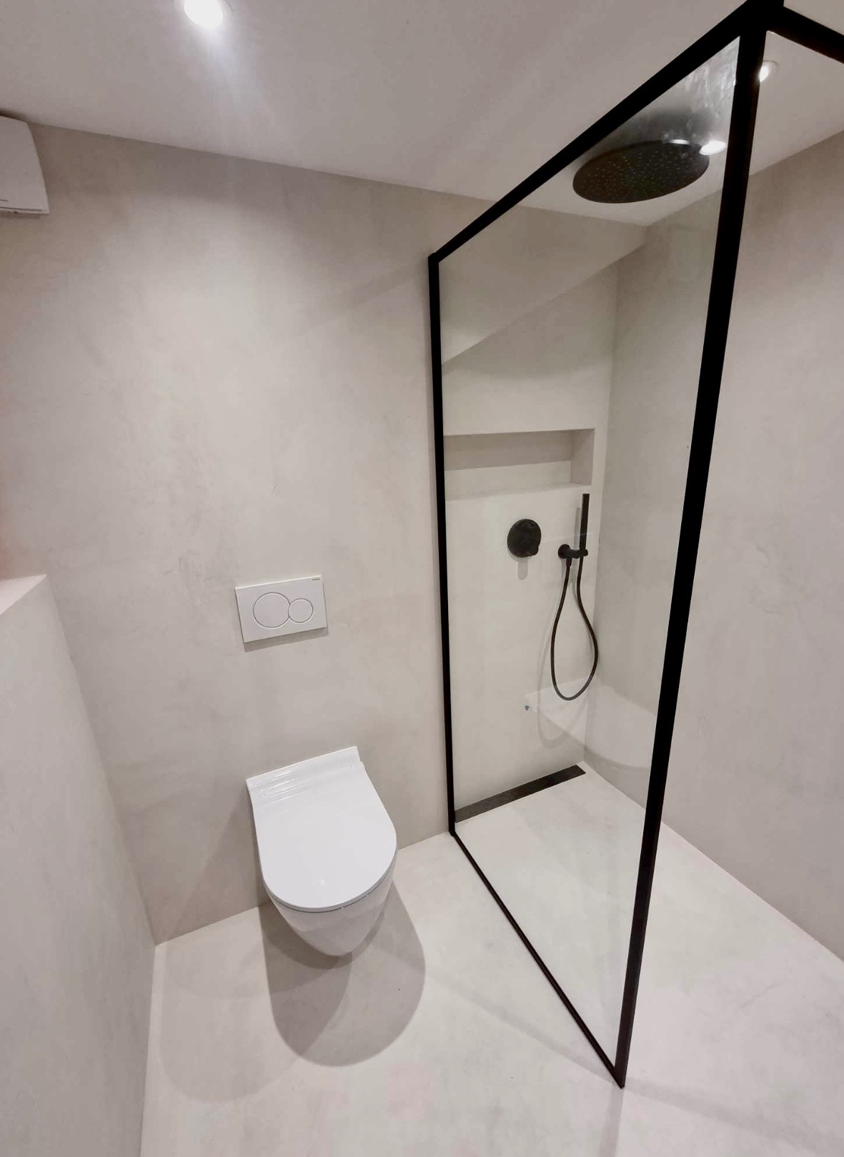 Minimalistyczna łazienka z kabiną prysznicową w czarnej ramie, wiszącą toaletą i ścianami wykończonymi w jasnym, matowym odcieniu.