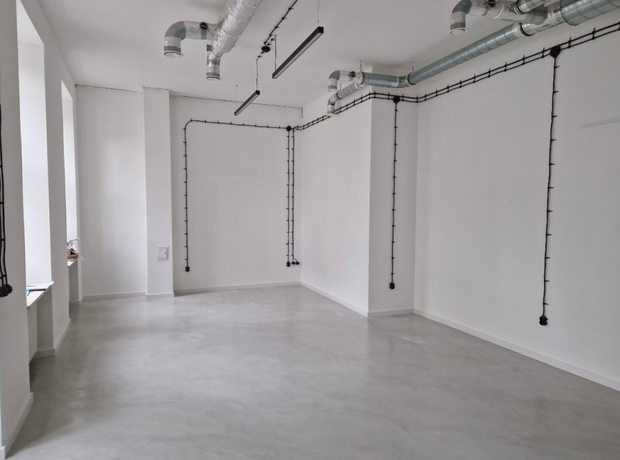 Jasnoszara, gładka posadzka w pustym, białym pomieszczeniu z widocznymi instalacjami elektrycznymi i wentylacyjnymi pod sufitem.