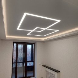 Ministerstwo sufitow - Nowoczesny pokój z białym sufitem, na którym umieszczono geometryczne oświetlenie LED w kształcie dwóch przecinających się kwadratów, widok na przeszklone drzwi balkonowe i grzejnik pod oknem.